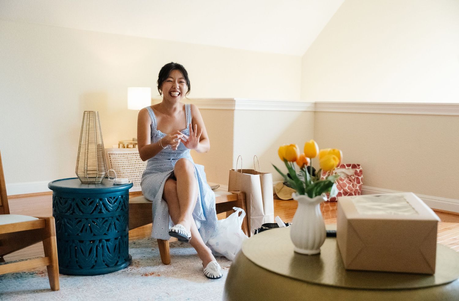 Happy bride in blue dress sits in bright hotel suite with yellow tulips nearby.