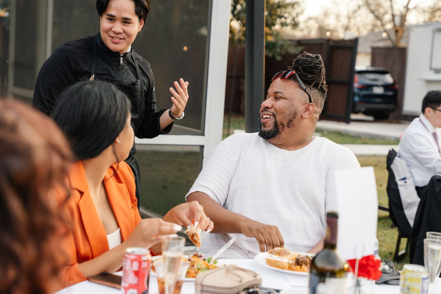 Guests laugh and enjoy food together at an outdoor dining table during a celebration.