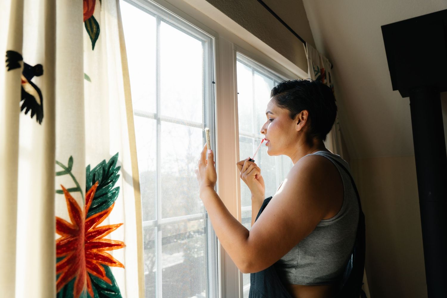 Young woman in gray top smiles while looking out a window with floral curtains.