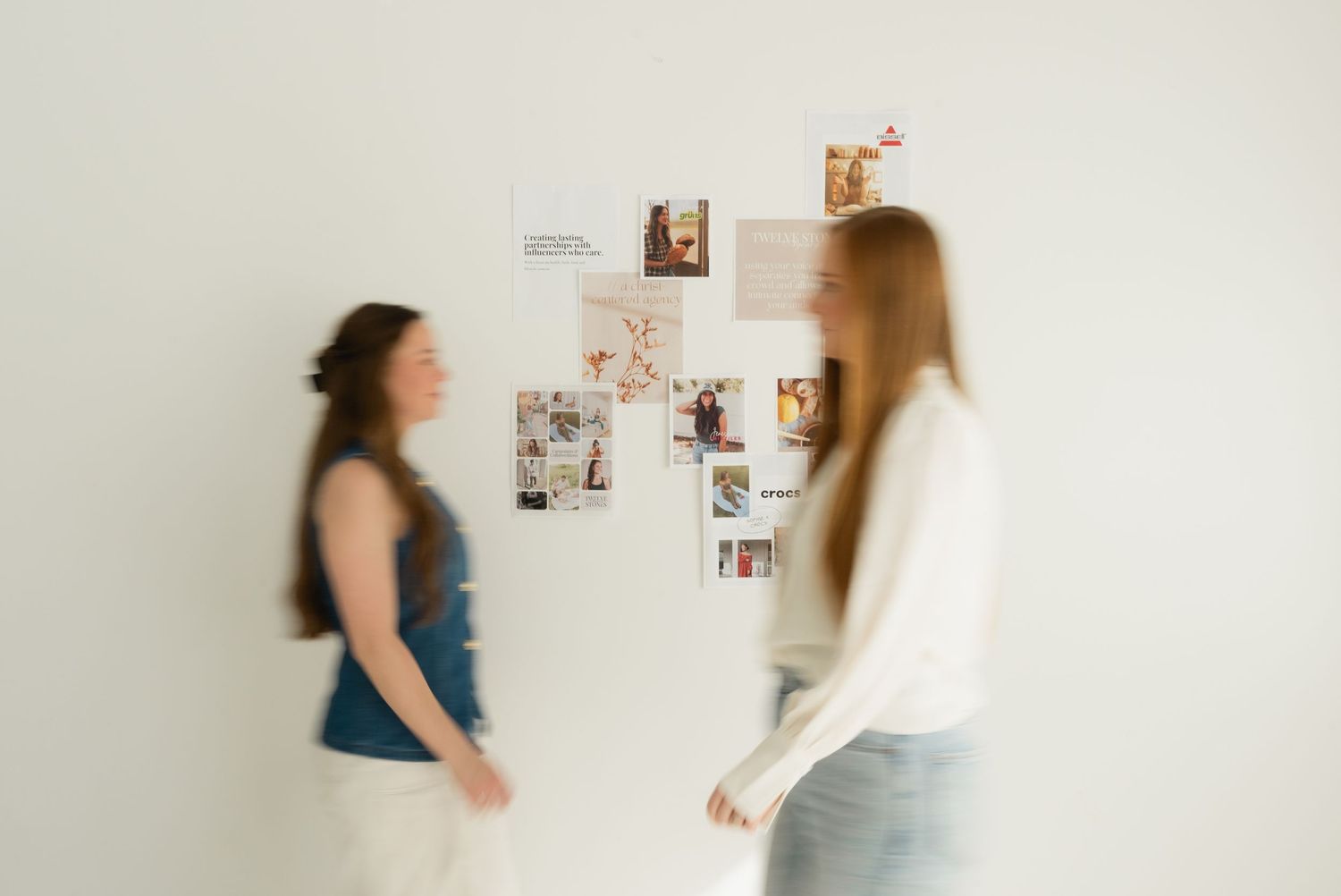 Two women in motion viewing a mood board of photos pinned to a white wall.