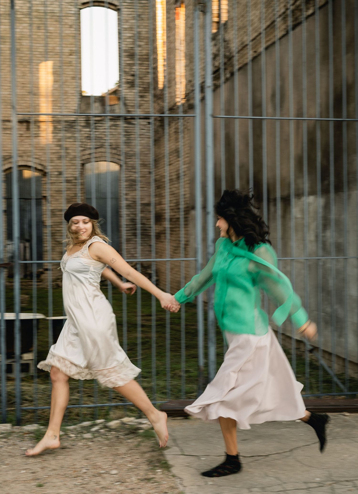 Two women in dresses holding hands and running through an urban courtyard.