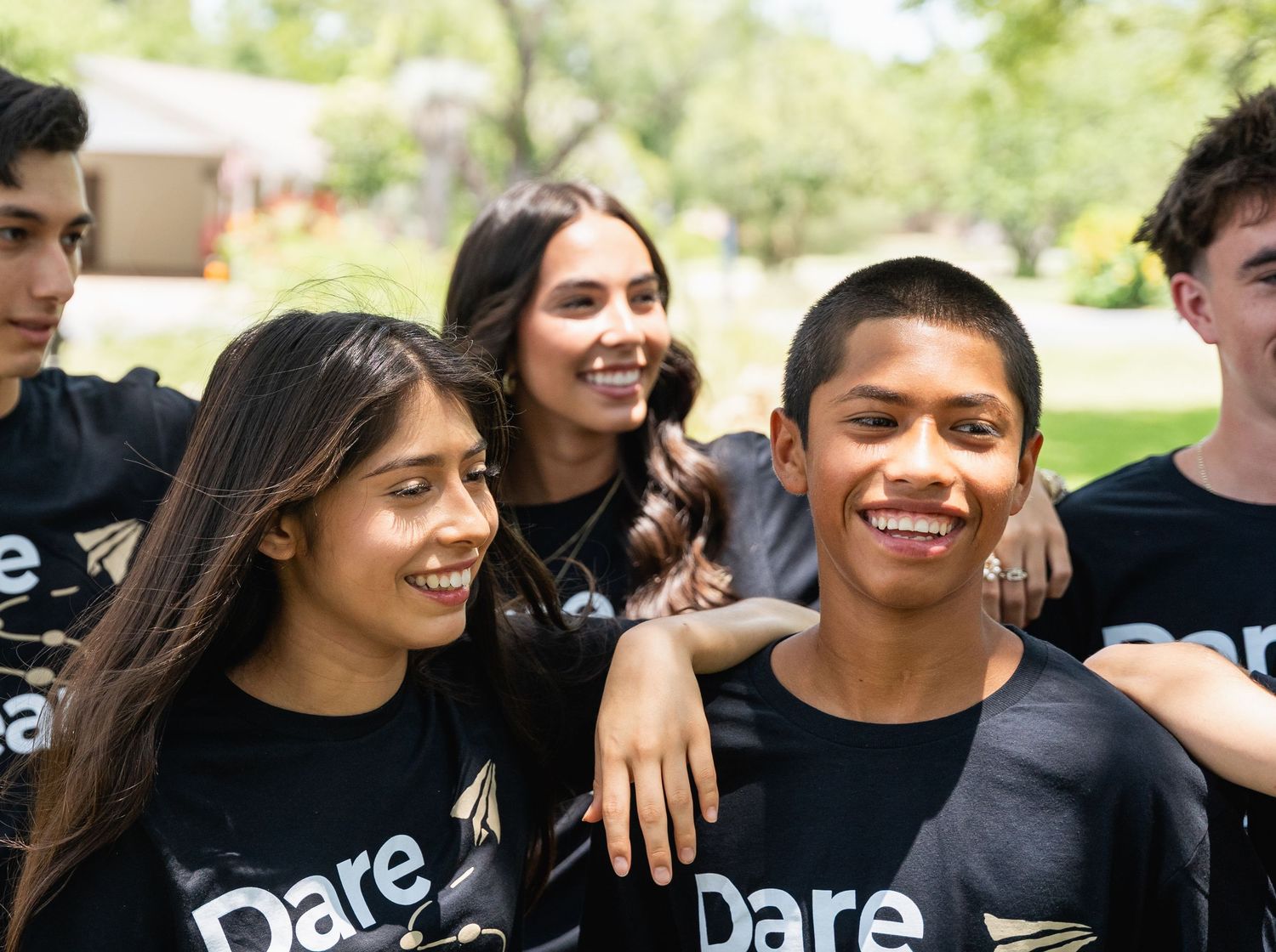 Group of smiling teenagers wearing black Dare shirts posing together outdoors in a park.