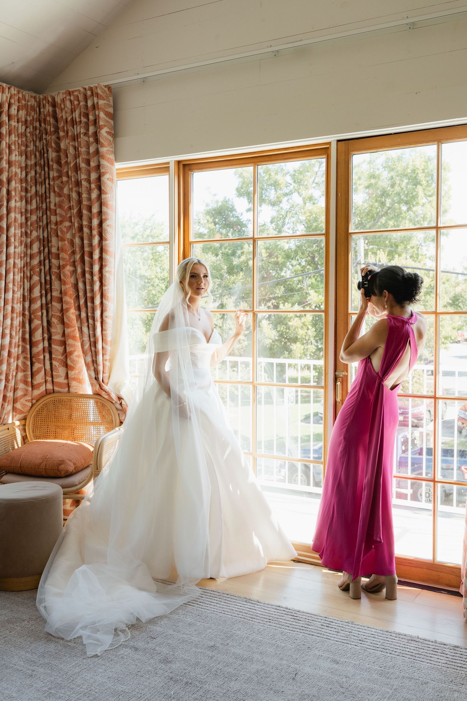 Blonde bride in flowing white ball gown with veil stands near large windows with bridesmaid in pink dress.