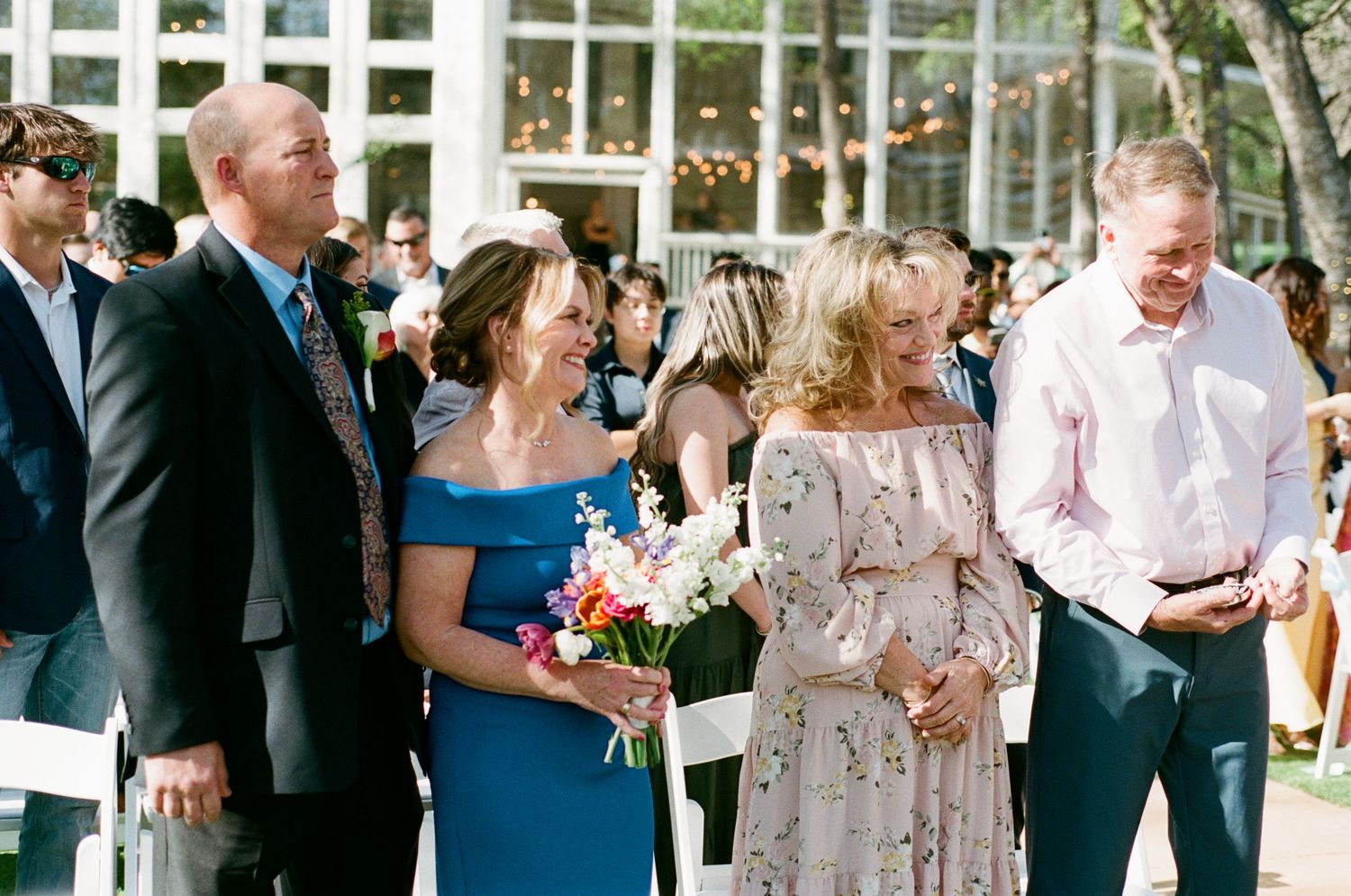 Wedding guests smiling and walking with colorful floral bouquet at an outdoor garden ceremony.