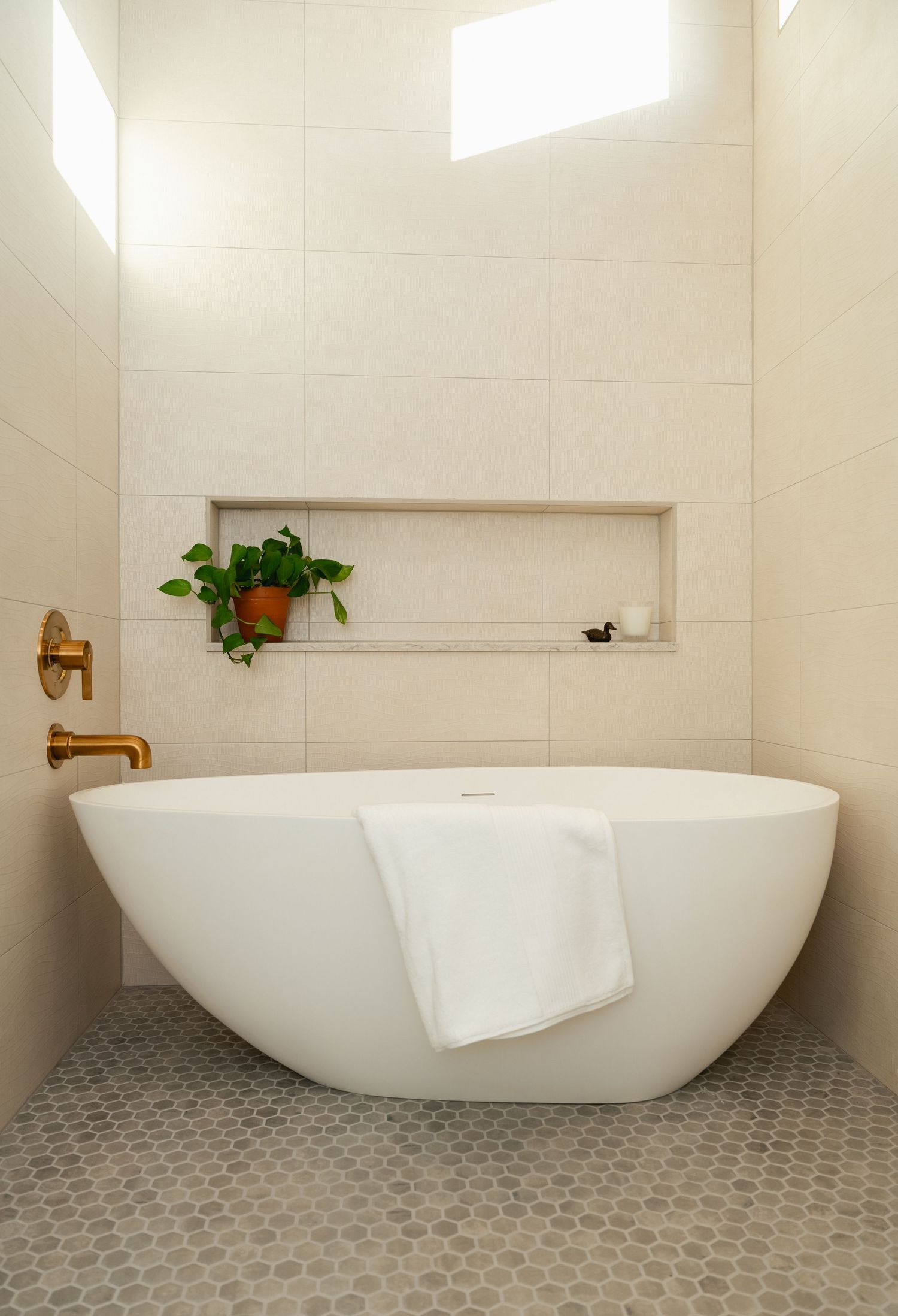 Minimalist freestanding soaking tub with penny tile floor, built-in niche, and brass wall-mount faucet.