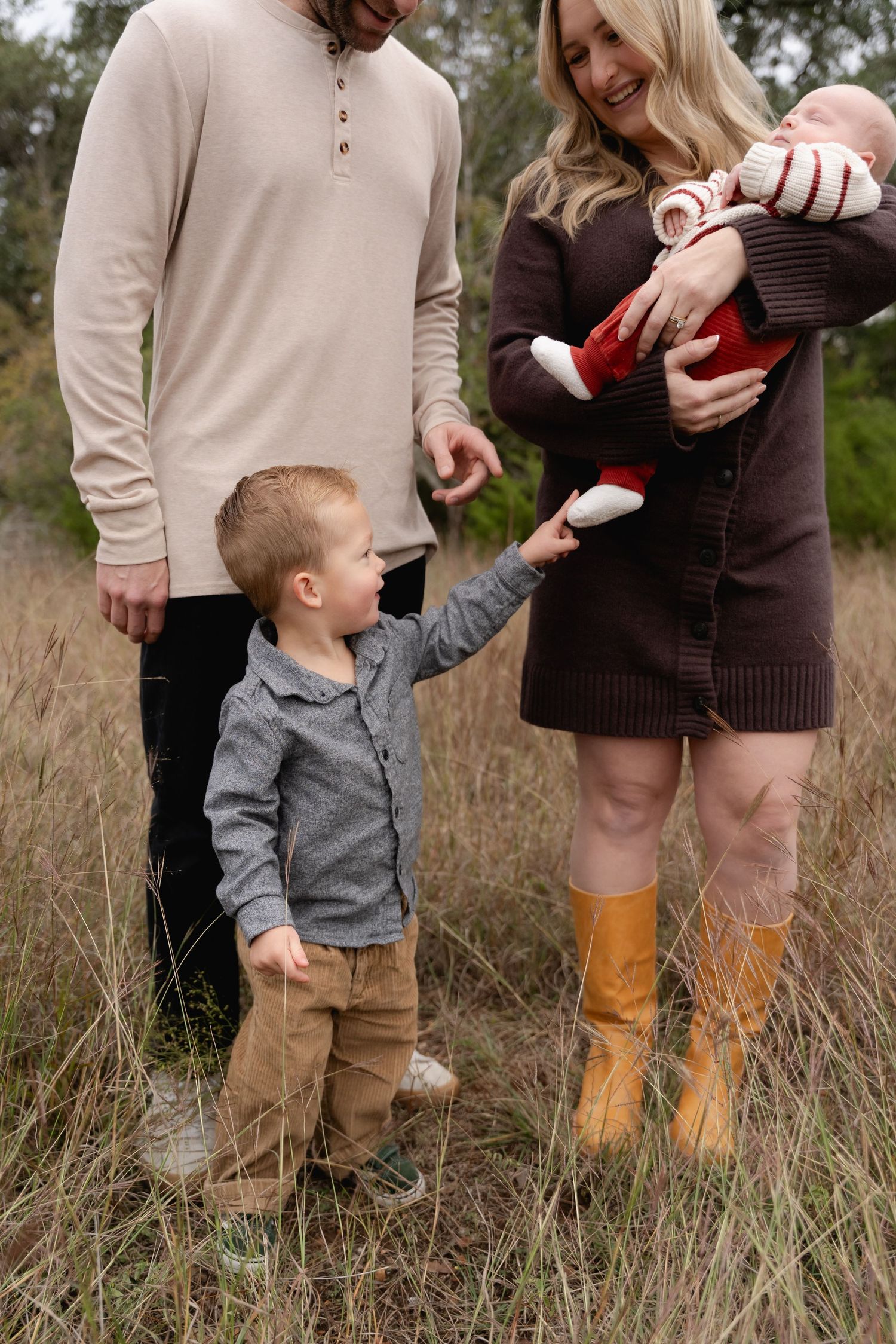 Family of four in a field, mother holding newborn, toddler son holding parents hands, fall outdoor photo session.