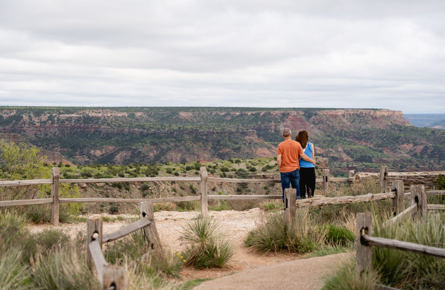 Two people stand arm-in-arm at a fenced overlook admiring vast canyon views under a cloudy sky.