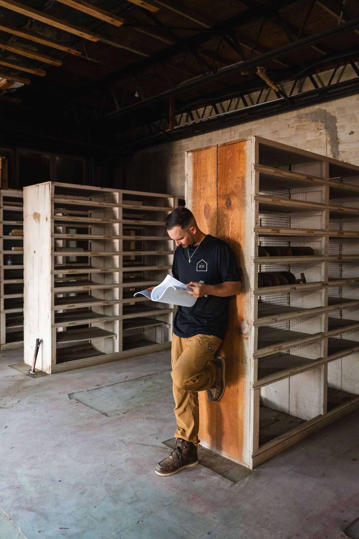A man reviews blueprints while leaning against wooden shelving in an unfinished construction space.