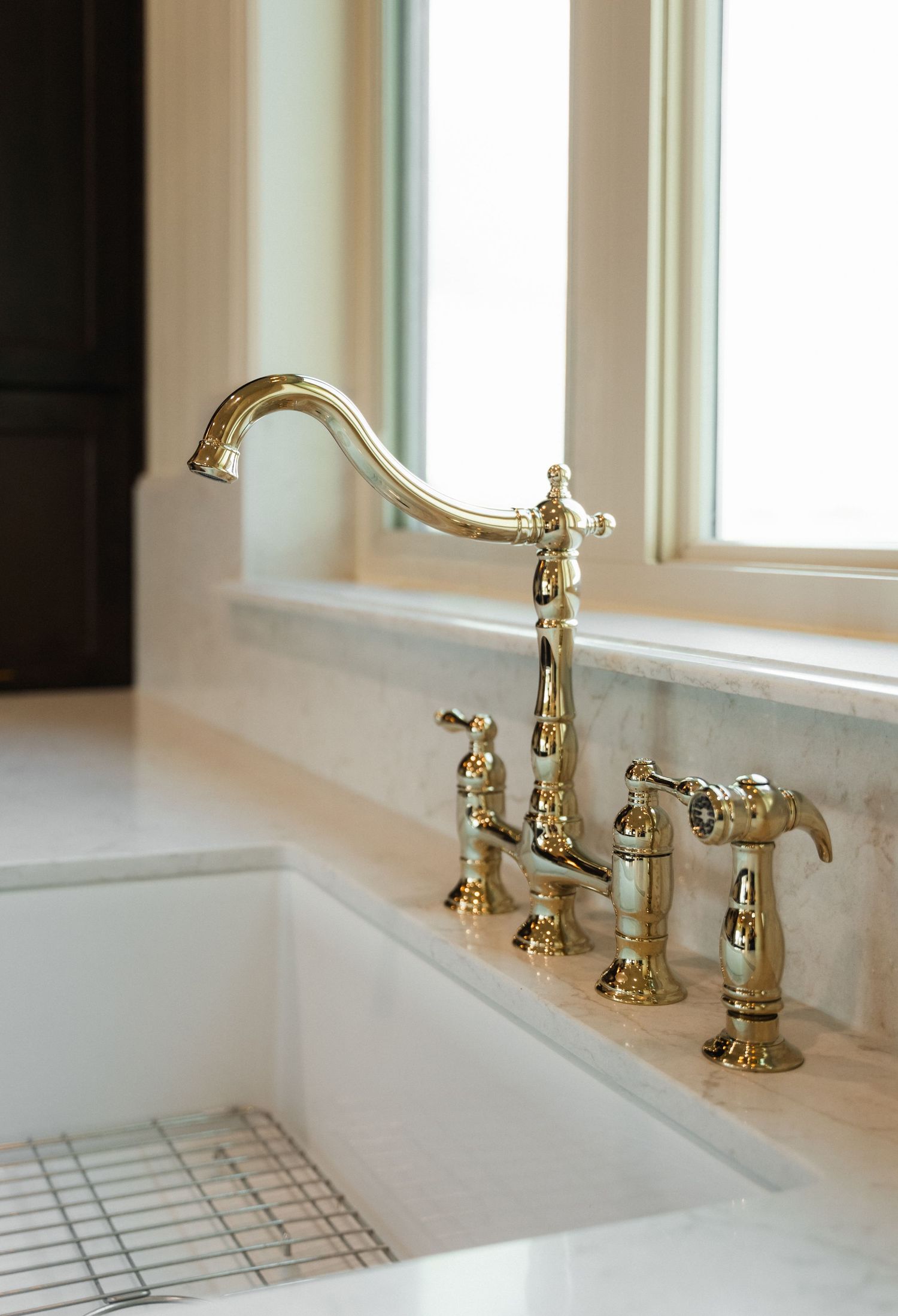 Elegant gold bridge kitchen faucet with sprayer on marble countertop near bright window.