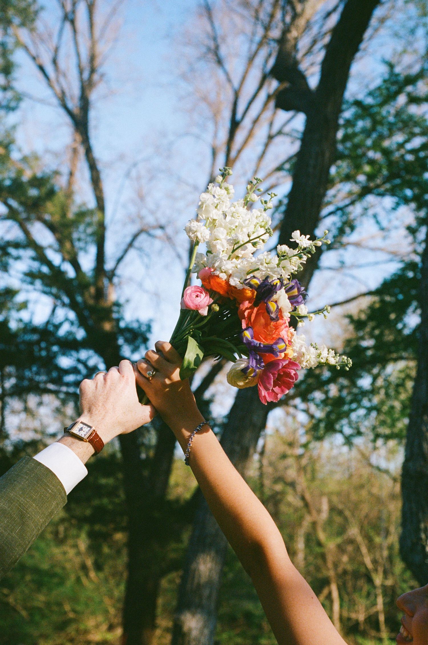 Two hands holding a colorful wedding bouquet of pink, white, and purple flowers aloft among bare trees outdoors.