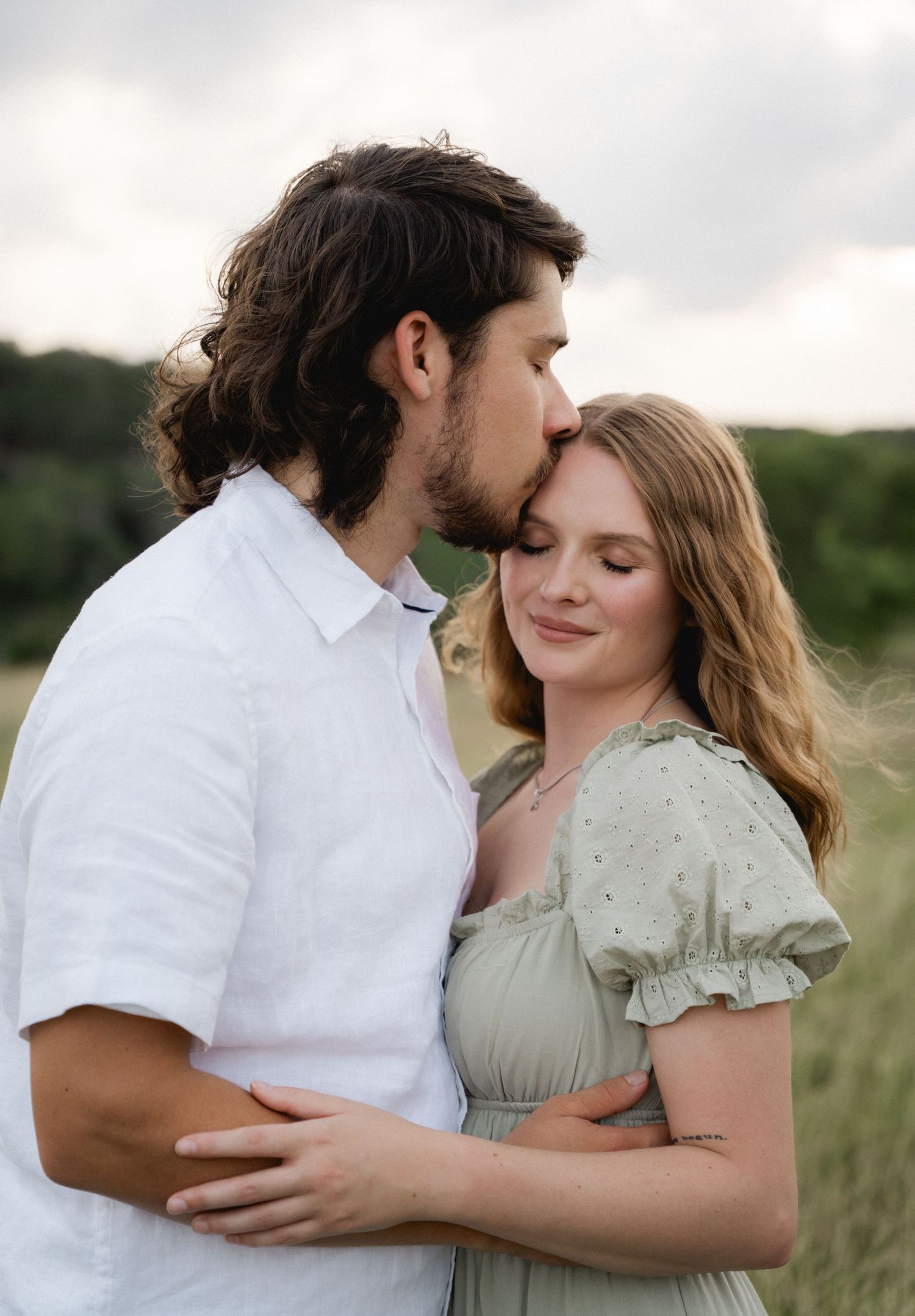 A couple shares a tender moment outdoors, man kissing woman's forehead in a green field at dusk.