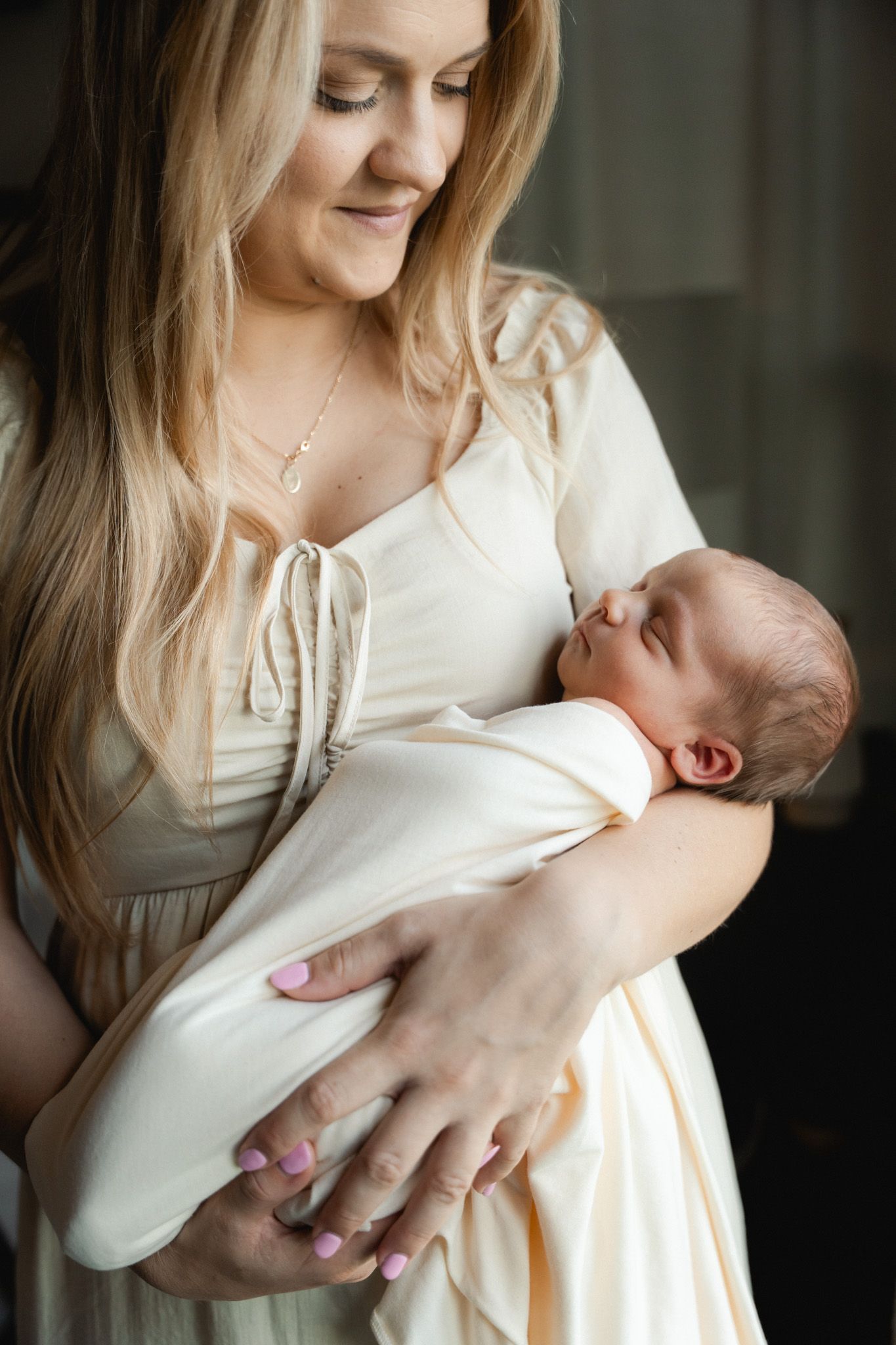 Smiling blonde mother in cream dress cradling swaddled newborn baby in her arms indoors.
