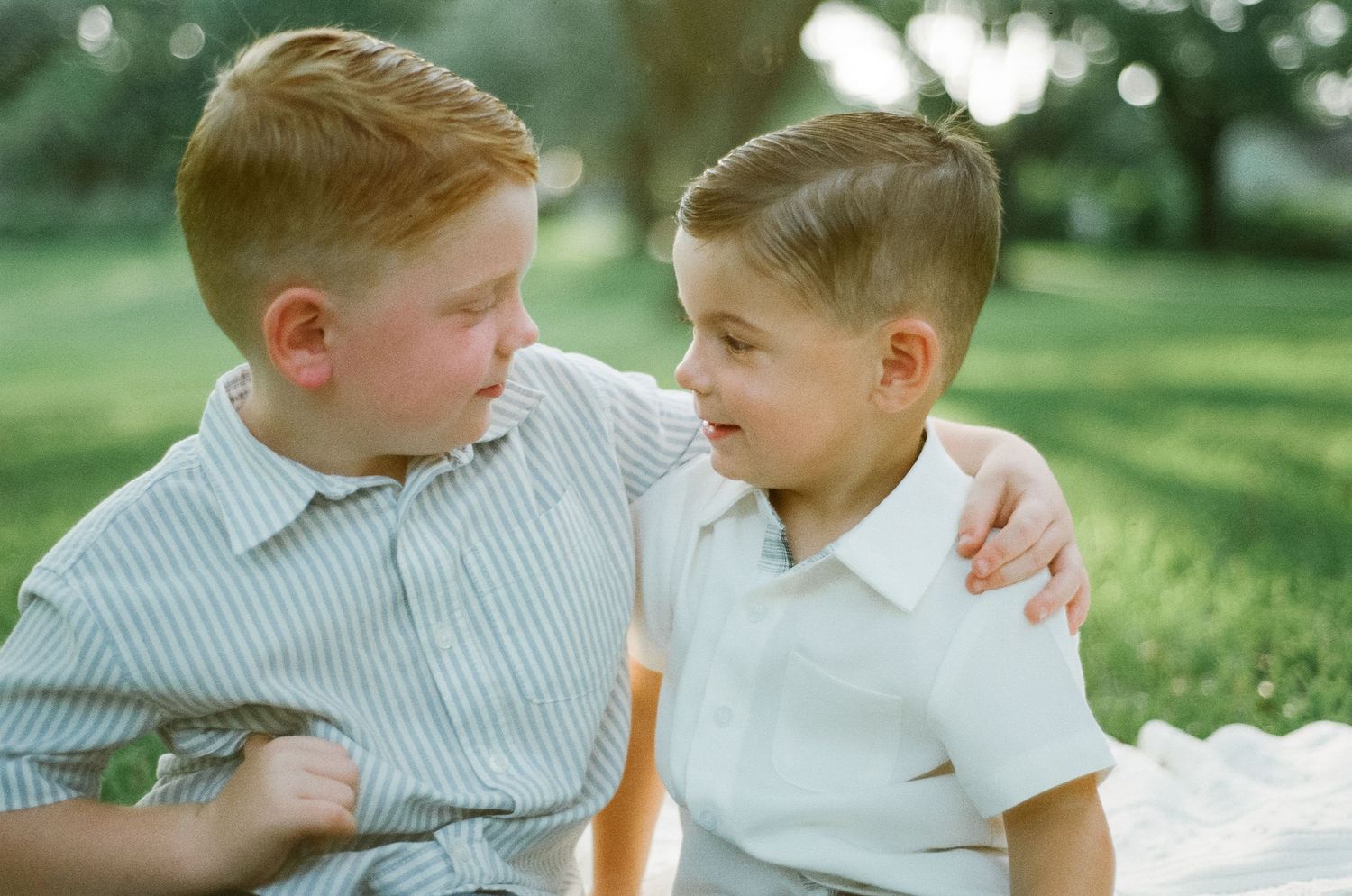 Two young red-haired boys sitting outdoors on grass, one with arm around the other, smiling at each other.
