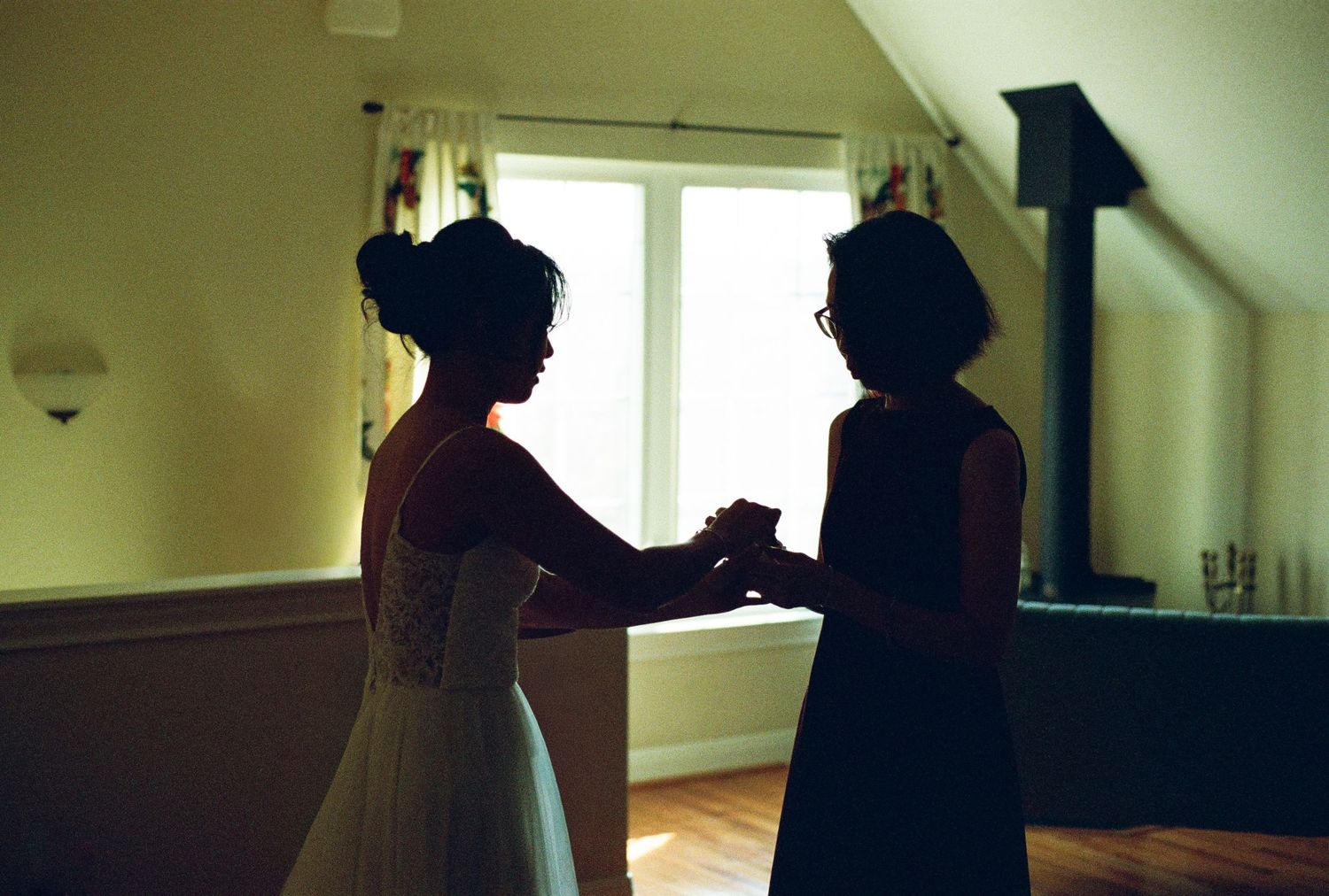 Bride and bridesmaid share tender moment holding hands in silhouette by bright window.