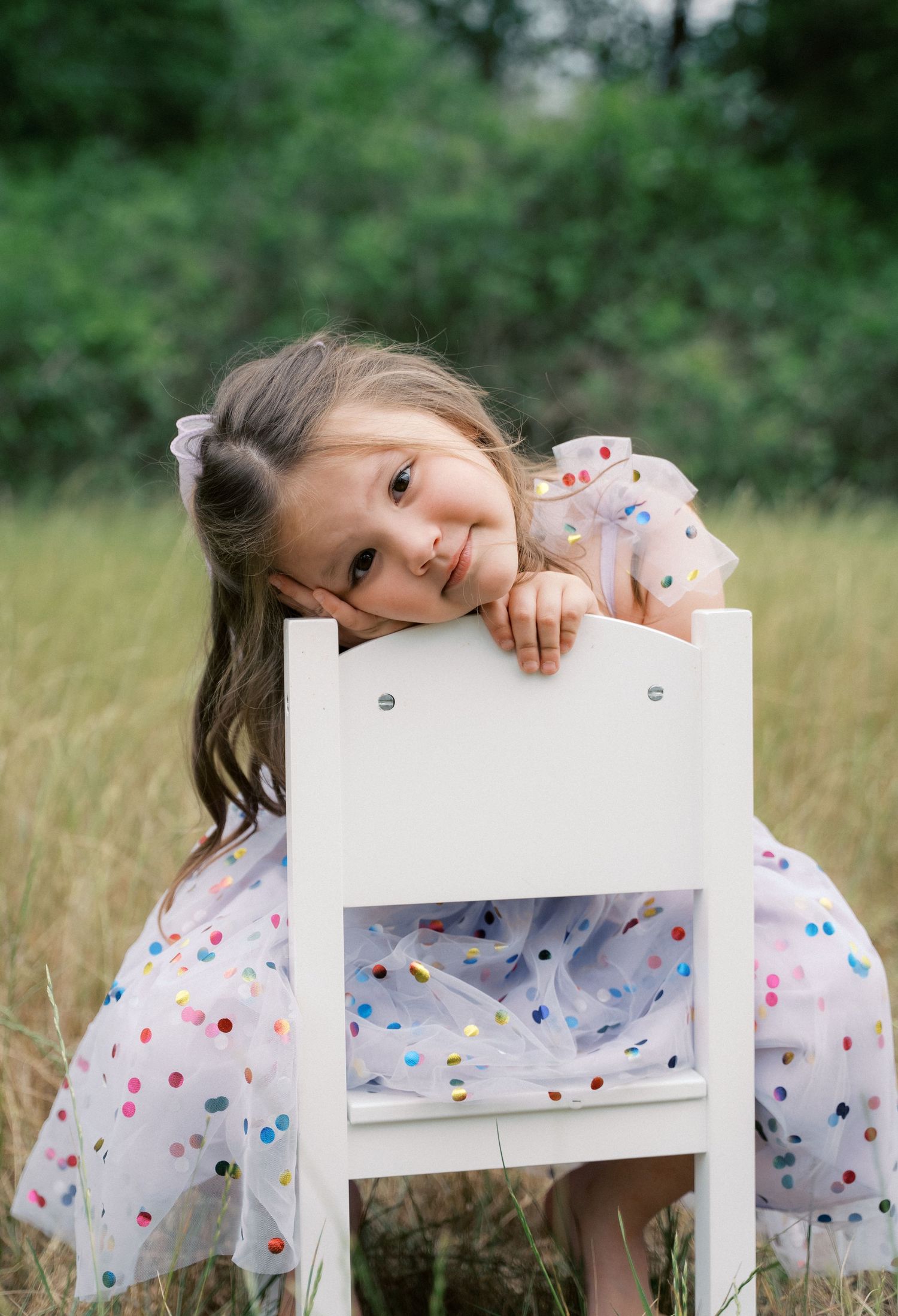 A young girl in a colorful polka dot dress rests her chin on a small white chair in a grassy field.