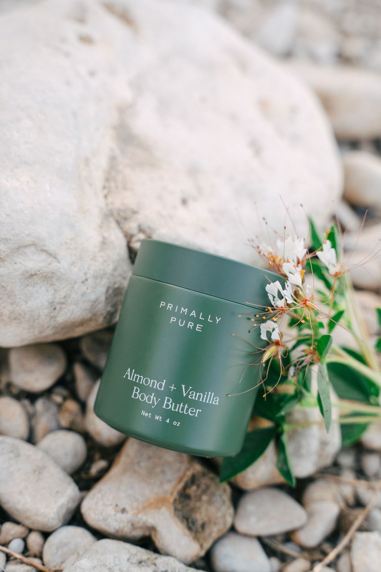 Primally Pure Almond and Vanilla Body Butter green jar on rocks with white wildflowers nearby.