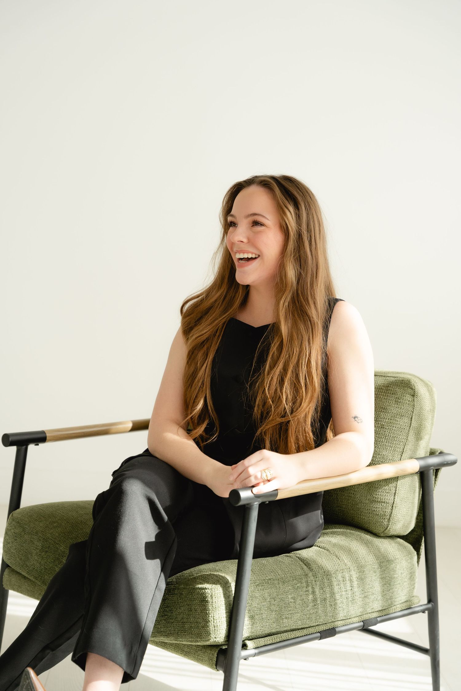 Laughing woman in black sleeveless outfit relaxing in a green velvet armchair in bright studio.