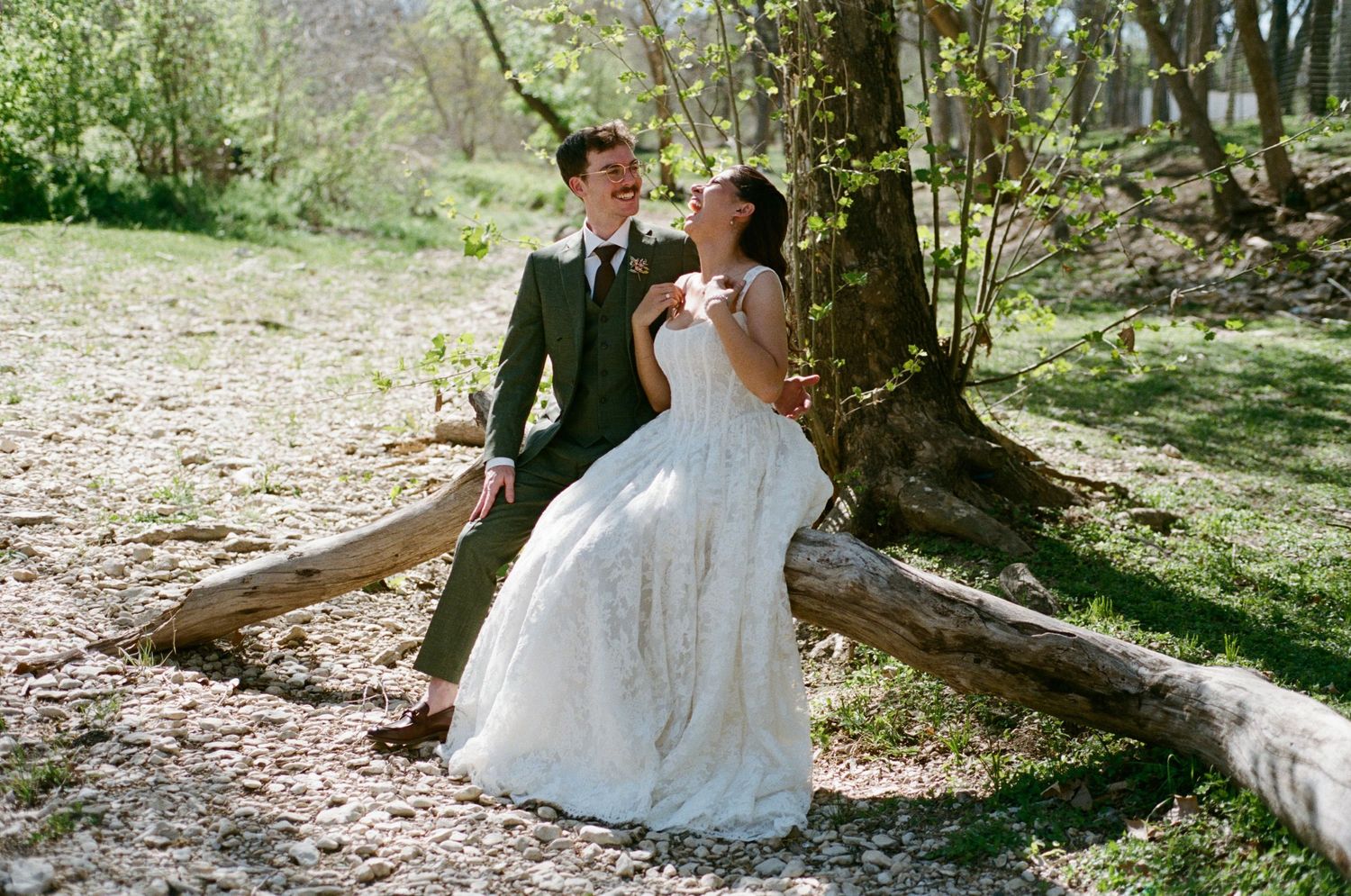 Bride and groom sitting together on a fallen log laughing in a sunlit outdoor forest setting.