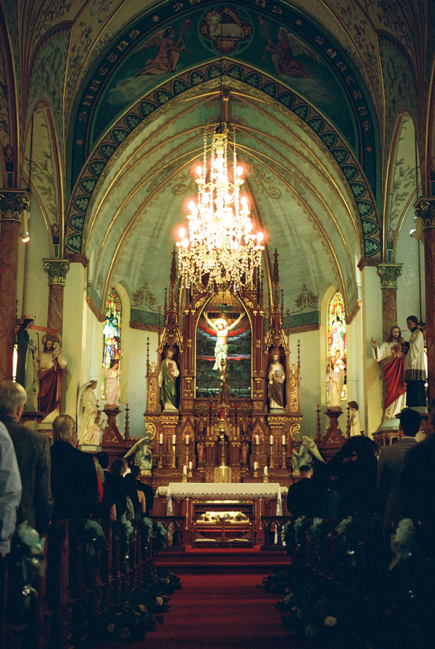 Ornate Catholic church interior with gilded altar, crystal chandelier, arched ceiling murals during wedding ceremony.