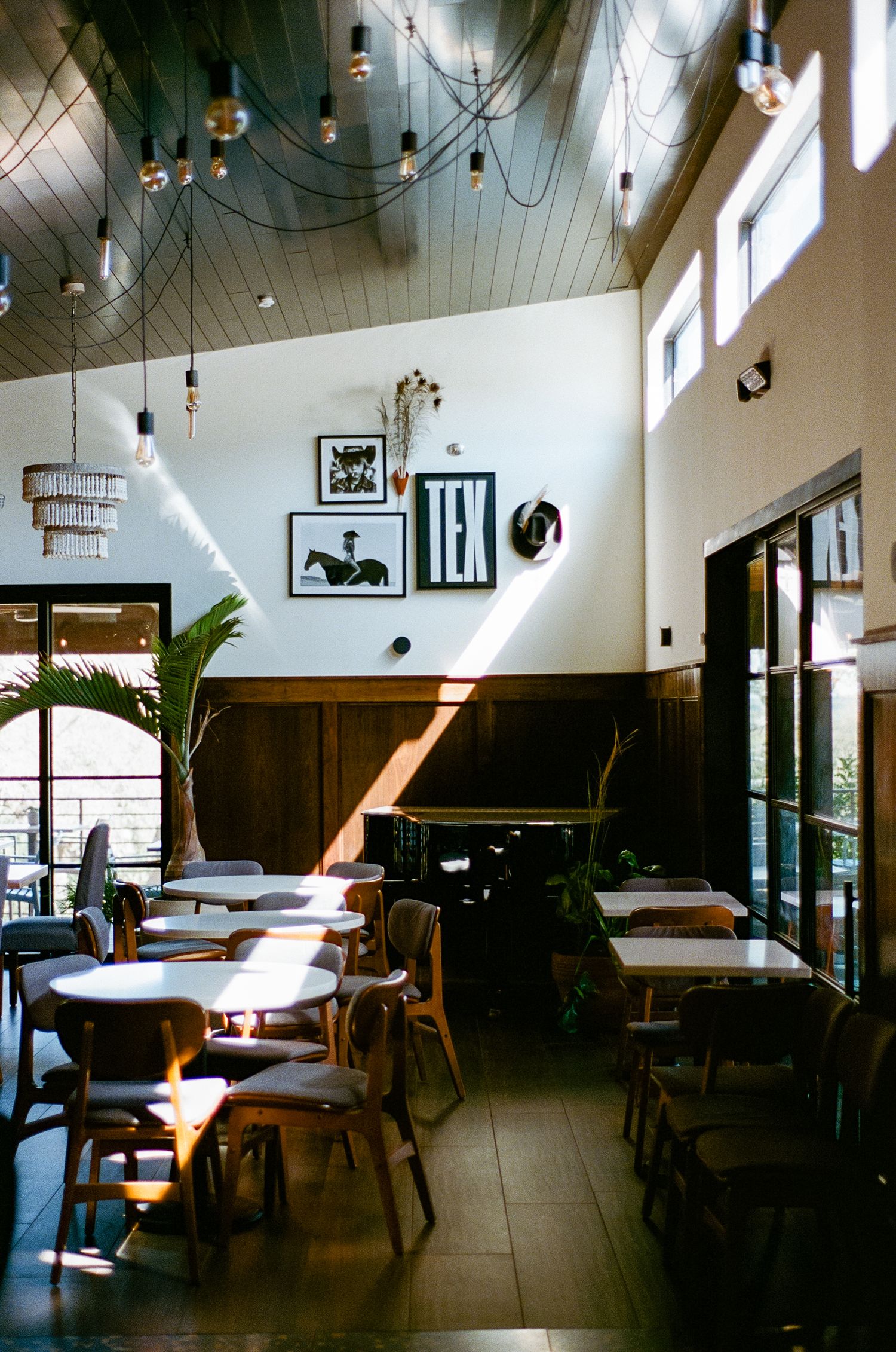 Sunlit restaurant with high vaulted ceilings, Edison bulb lighting, wooden chairs, and TEX wall art decor.