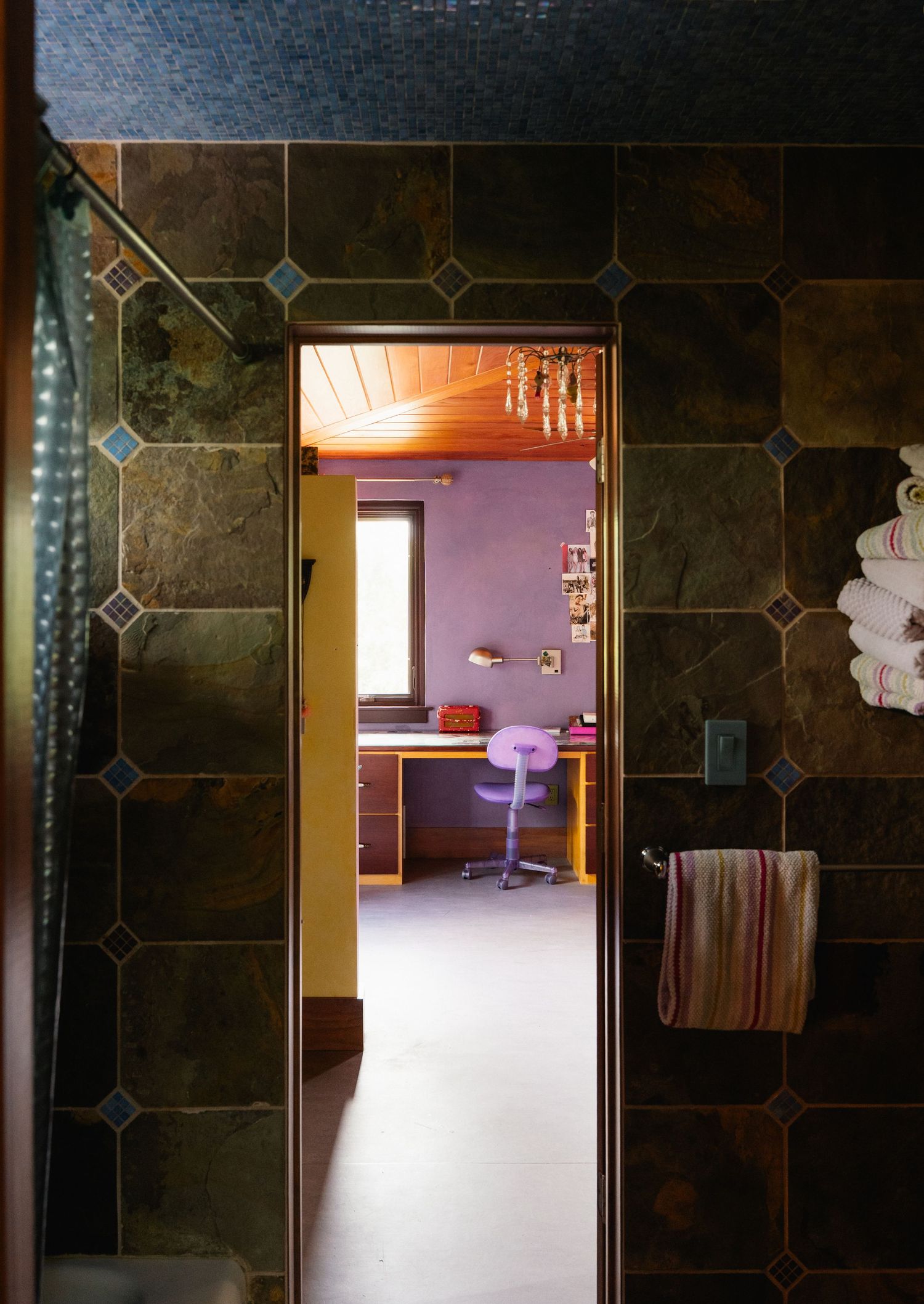 Dark slate tile bathroom with doorway leading to purple bedroom with desk and chair visible through entrance.