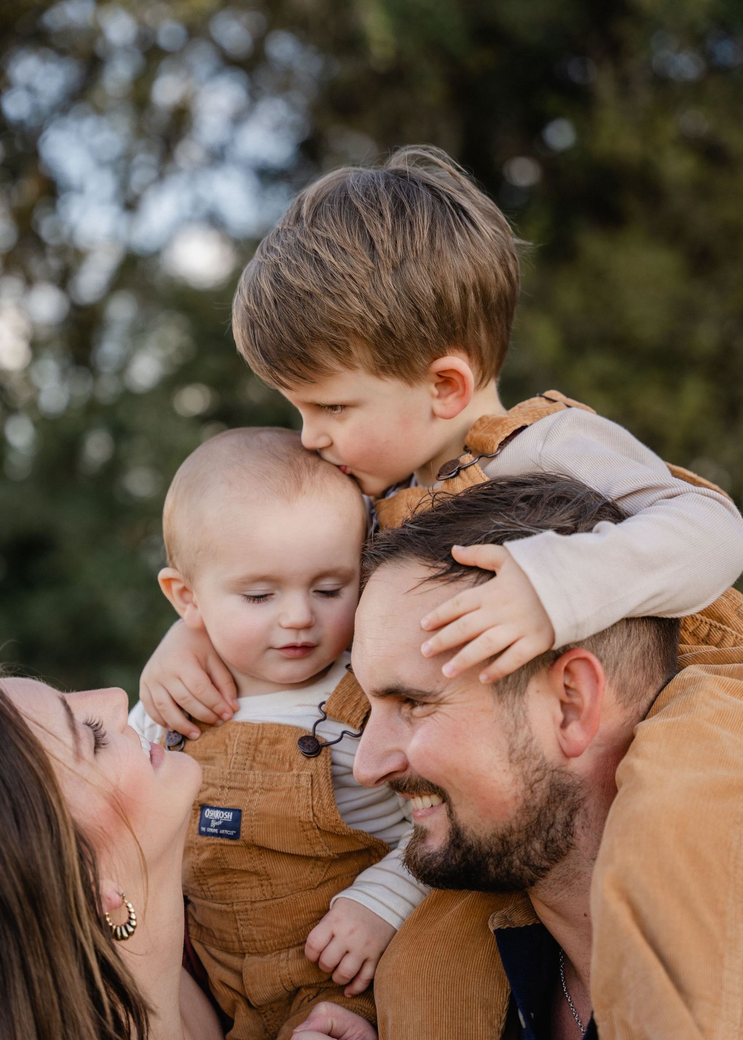 Parents and two young sons sharing a tender moment outdoors, older boy kissing babys head.