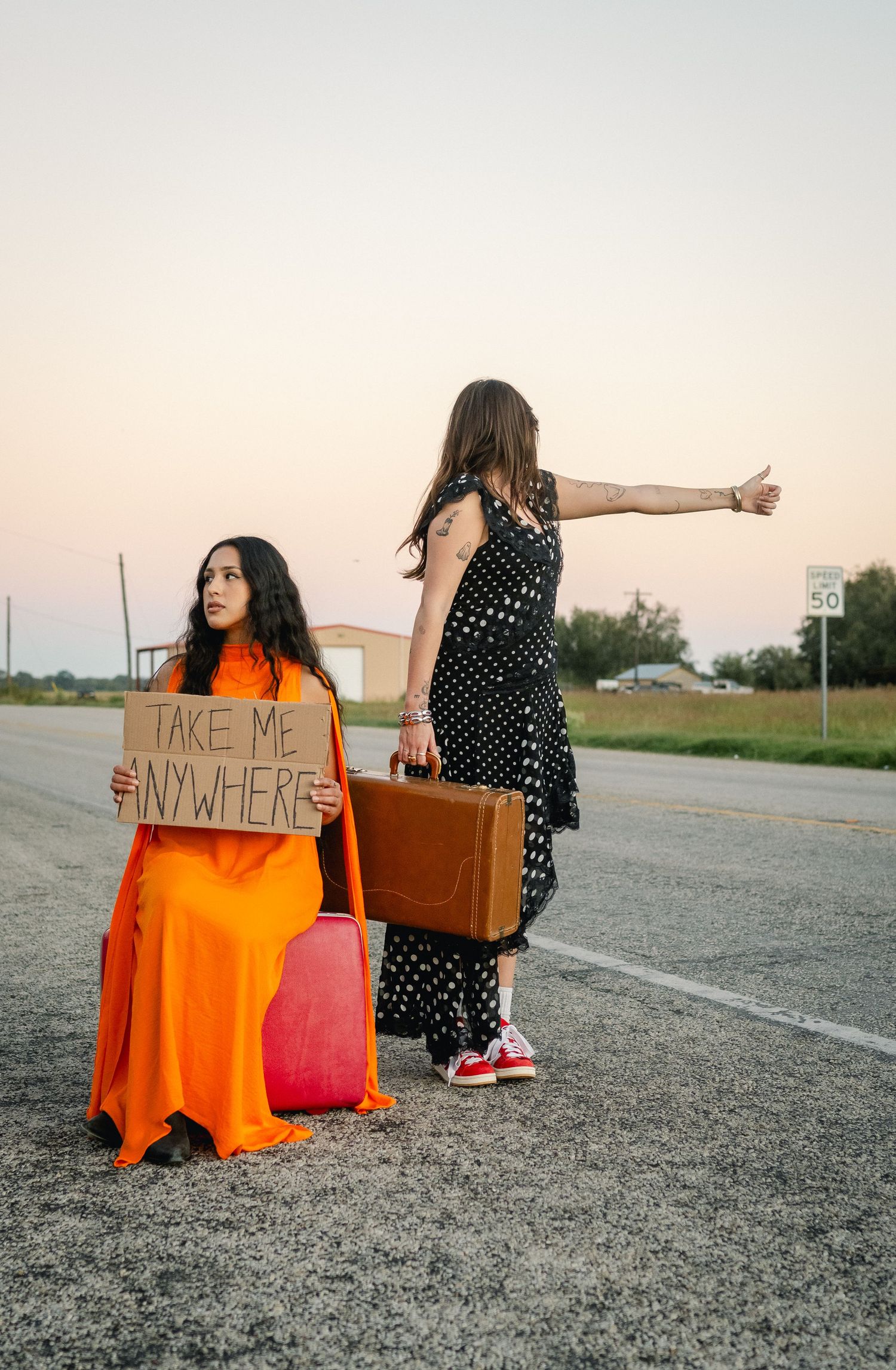 Two women hitchhiking on a roadside, one holding a cardboard sign reading Take Me Anywhere, with vintage suitcases.