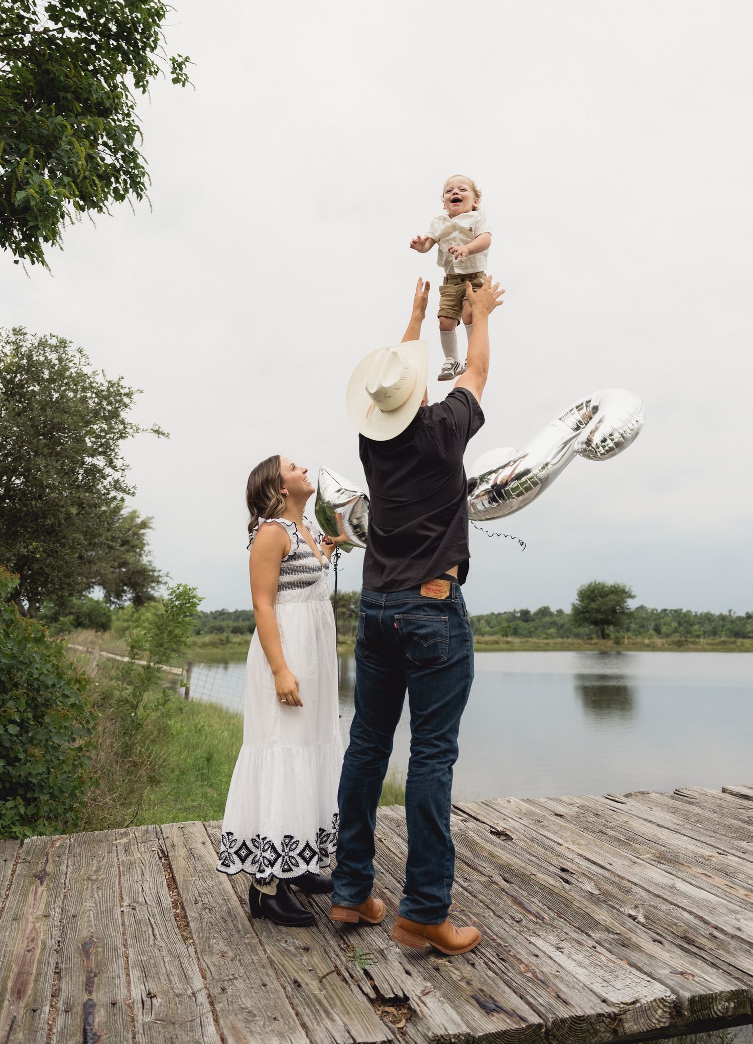A father in a cowboy hat tosses his laughing baby in the air on a lakeside dock.