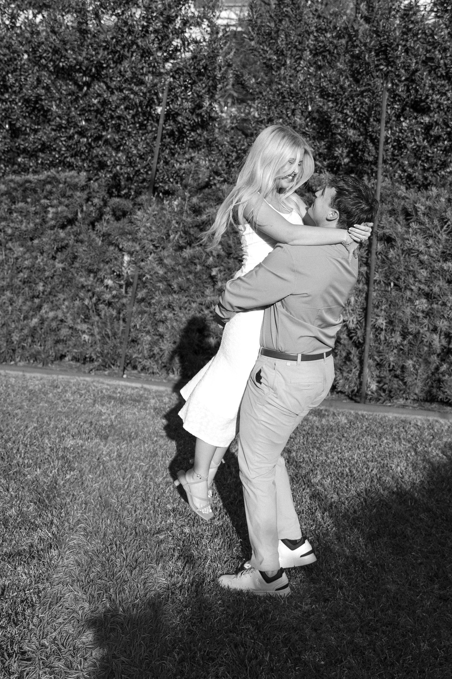 Black and white photo of man lifting woman in white dress outdoors, joyful romantic couple portrait.