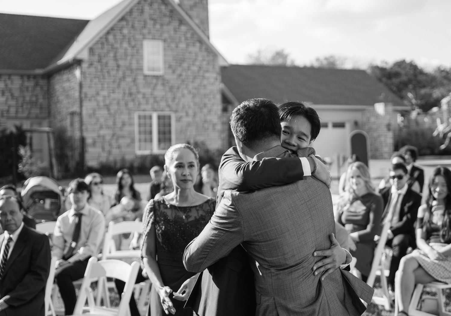 Emotional black and white photo of groom hugging family member at outdoor wedding ceremony.