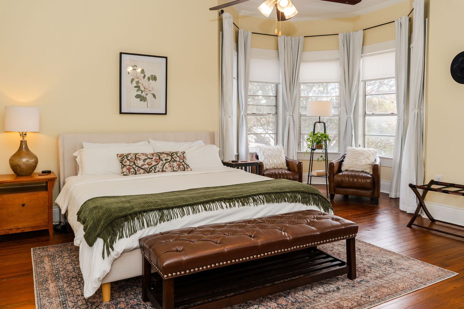 Elegant master bedroom with bay windows, upholstered bed, green throw blanket, leather bench, and hardwood floors.