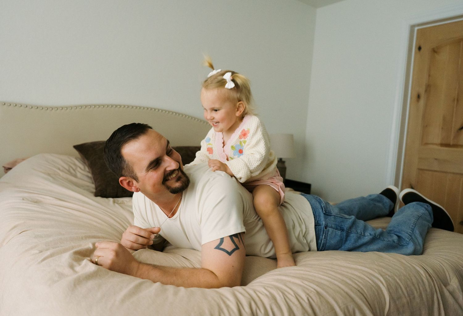 A father with tattoos playing with his young daughter on a beige bed.