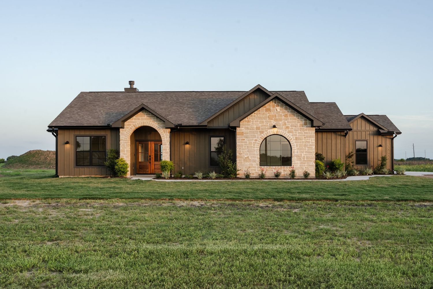 Ranch-style home exterior with stone and board-and-batten siding, arched entry, and dark roof at dusk.