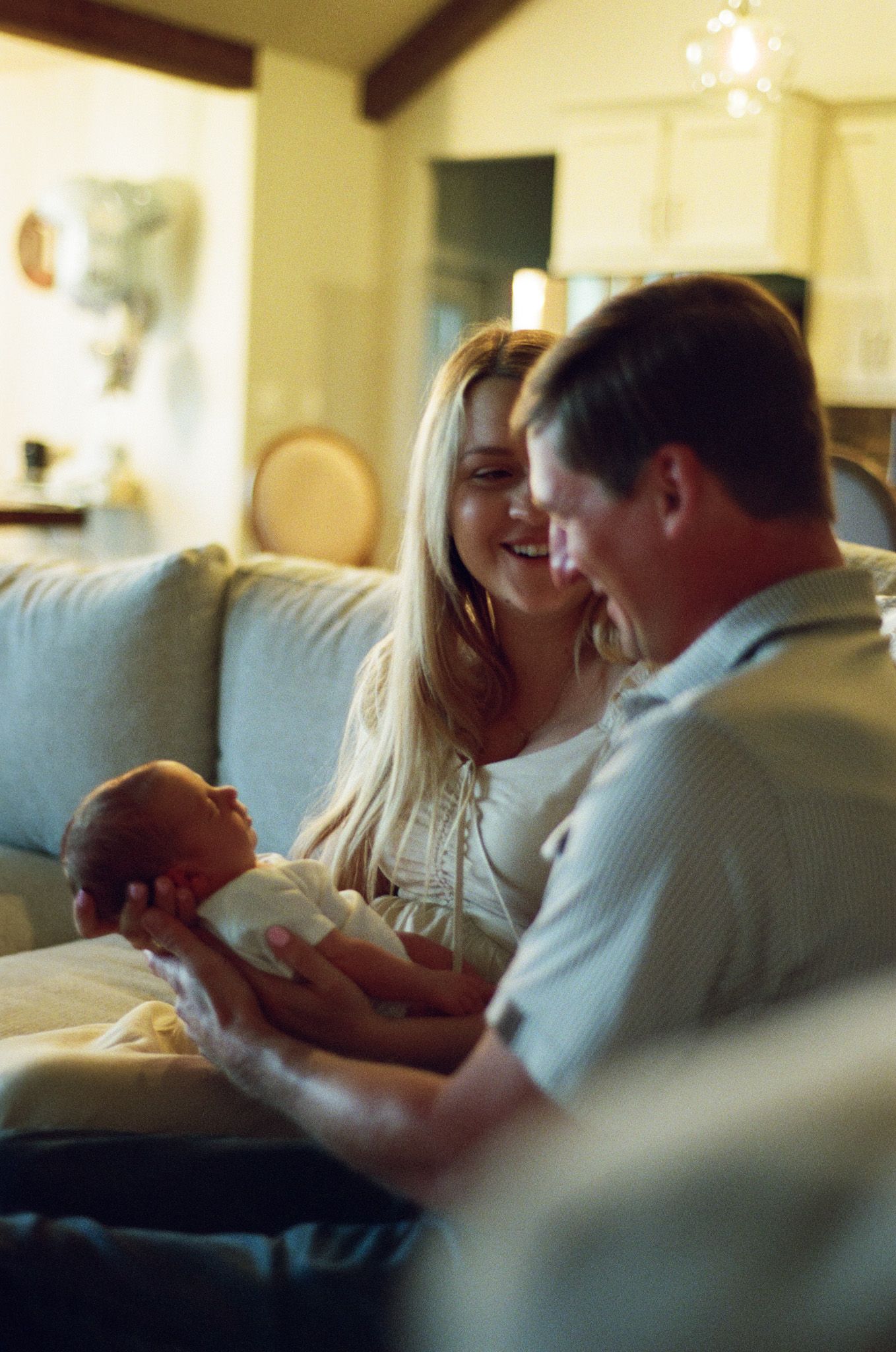 A smiling couple sitting on a couch together, lovingly holding their newborn baby in a warm, softly lit living room.