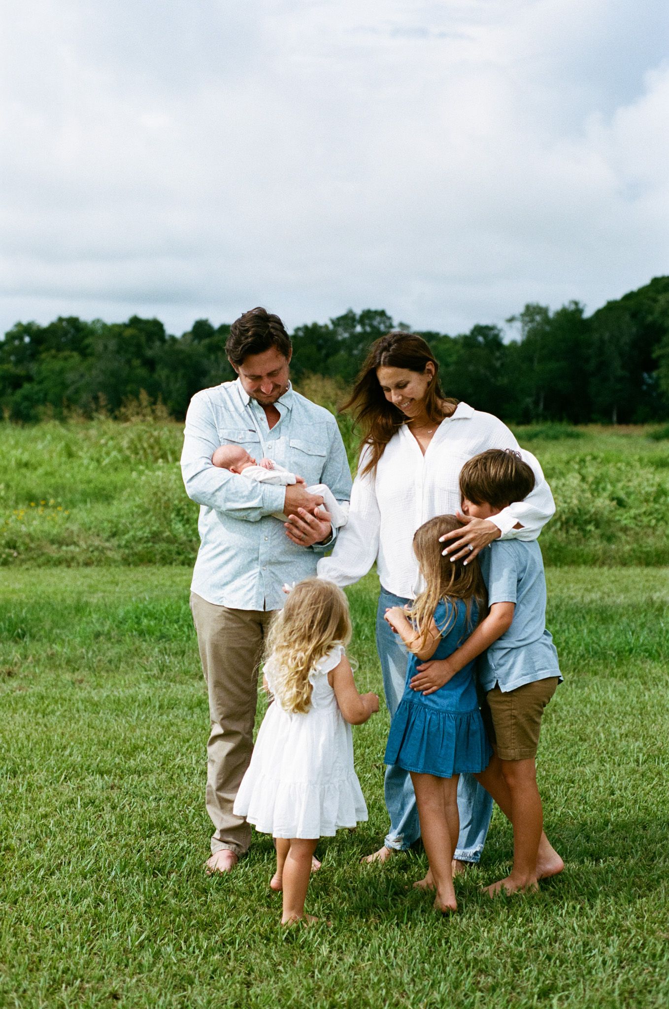Happy family of six stands in a green field, parents holding newborn baby.