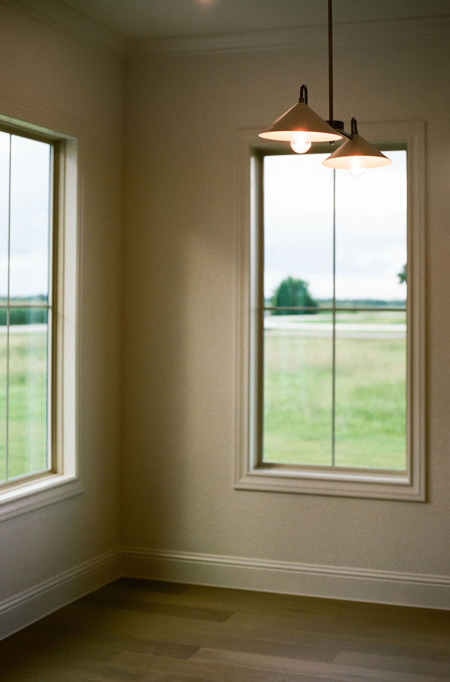 Bright corner room with two white-framed windows overlooking green fields, hardwood floors, and two pendant lights hanging from ceiling.