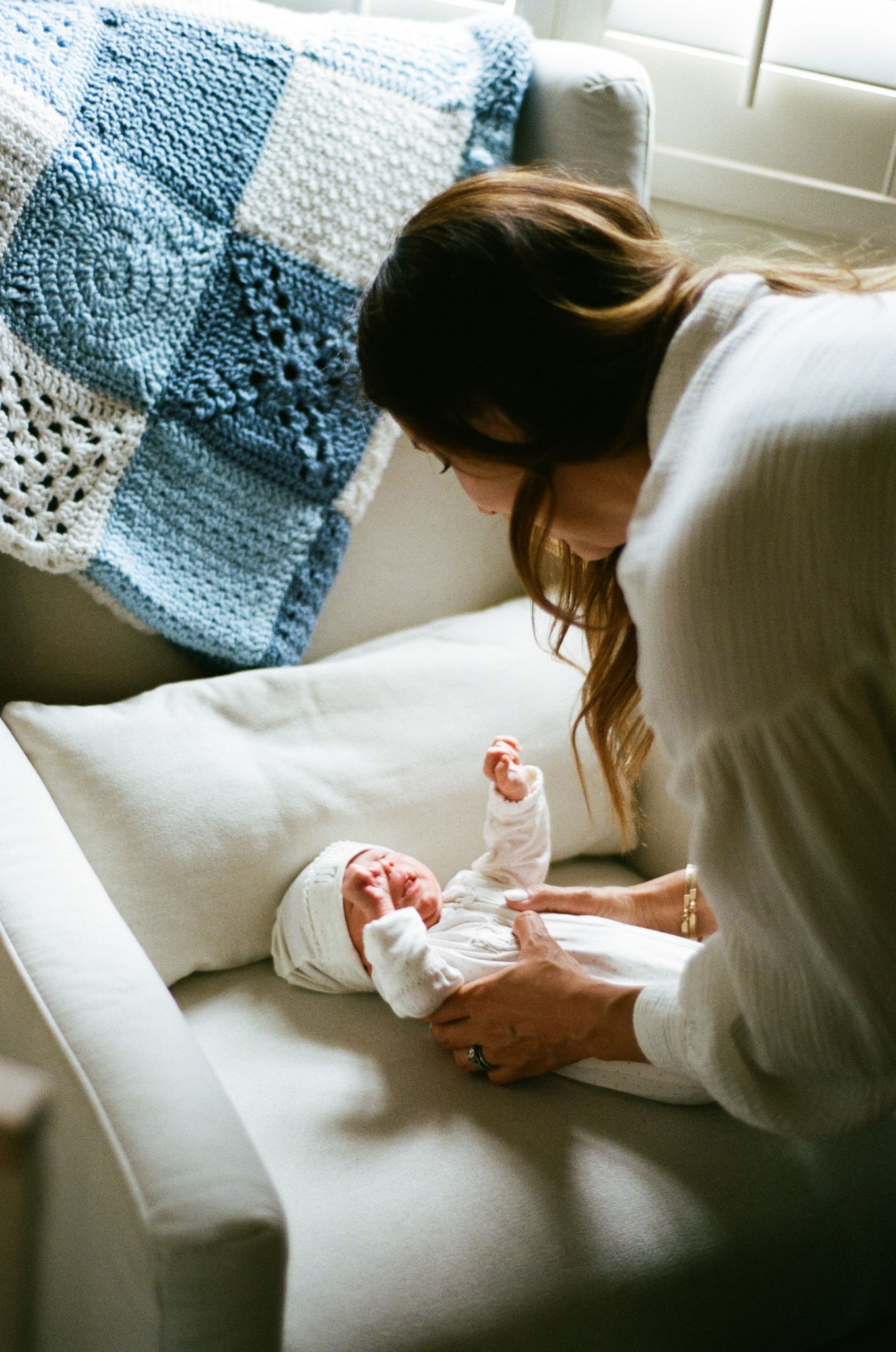 Mother gently caring for swaddled newborn baby on a cream armchair beside a blue crocheted blanket.