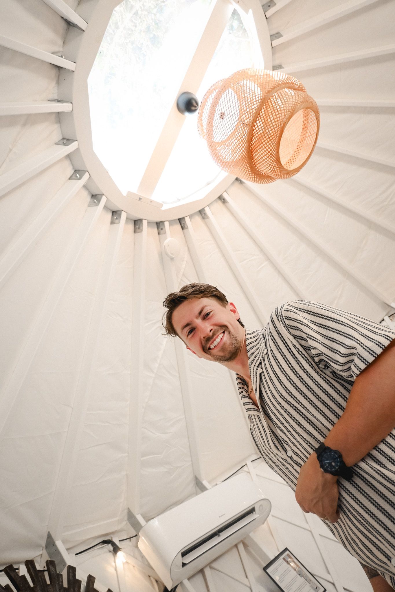 Man smiling inside a yurt with circular skylight, orange pendant fan, and white canvas walls.