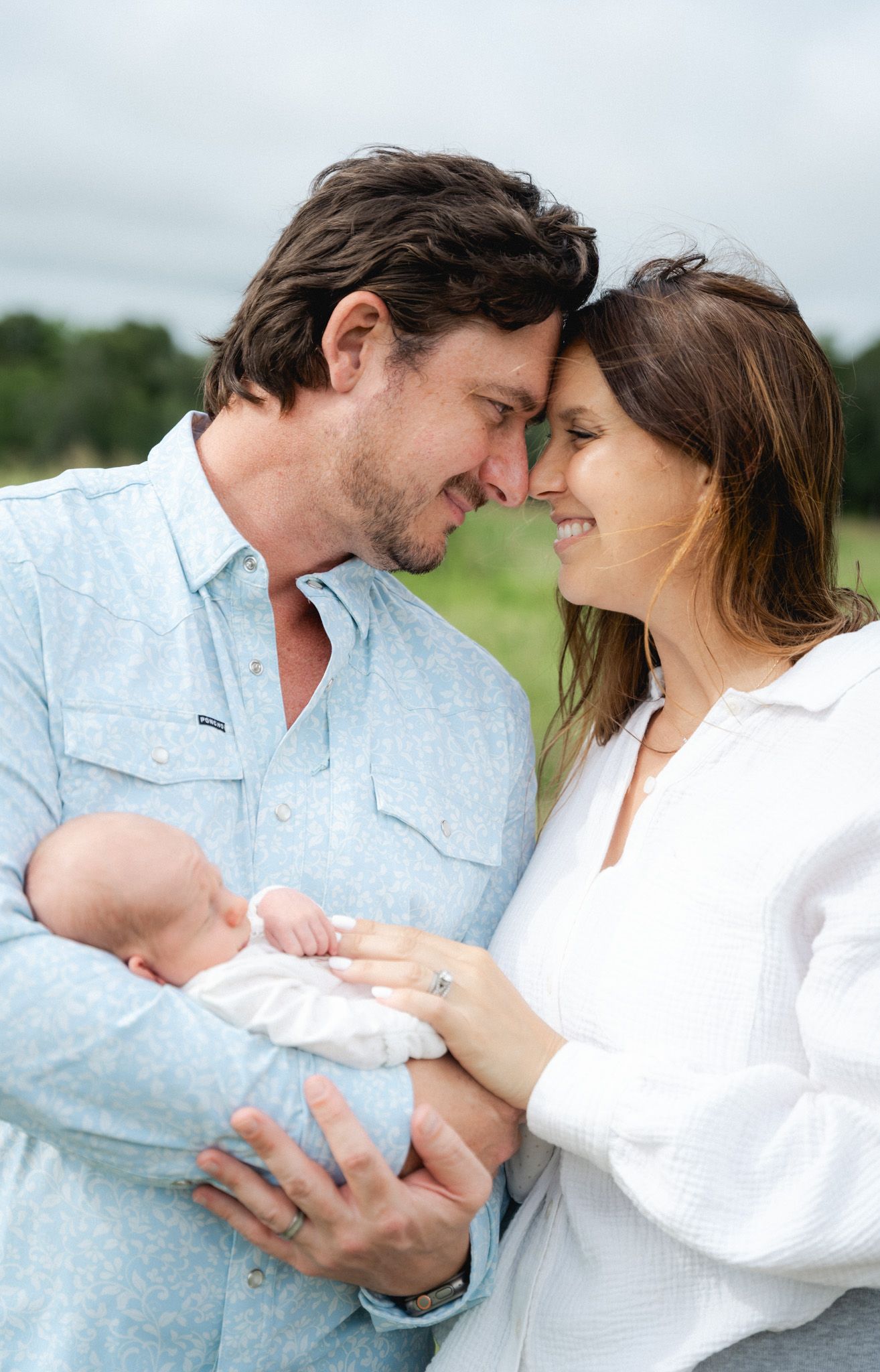 New parents touching foreheads tenderly while holding their newborn baby outdoors in a green field.