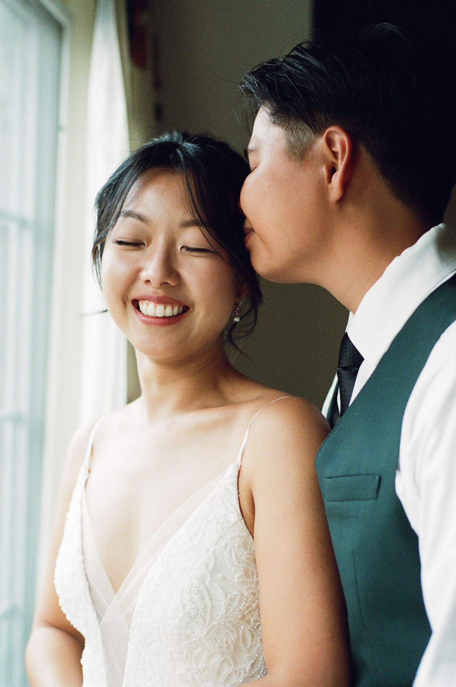 Groom kissing smiling bride on cheek in soft natural window light indoors.
