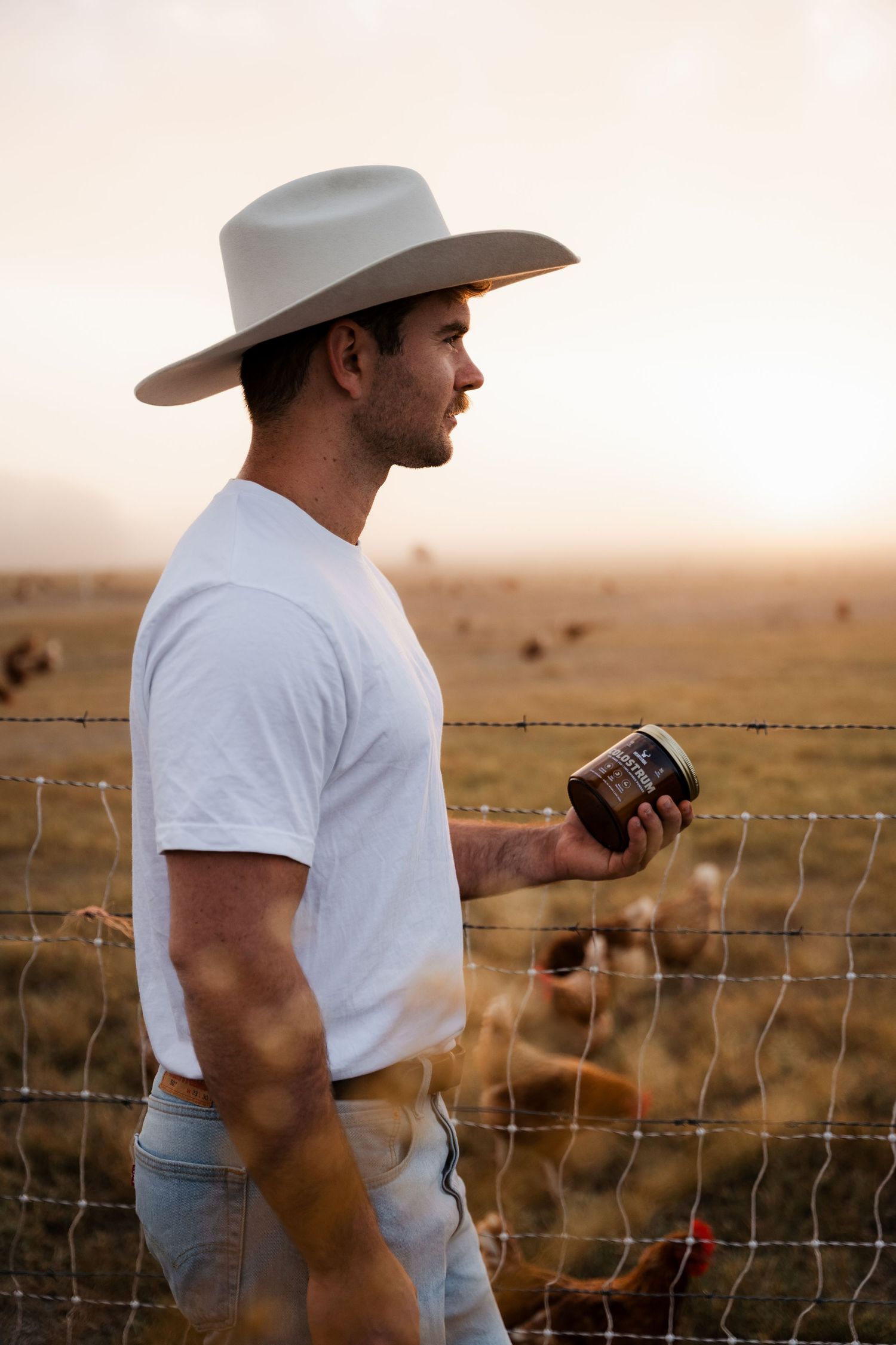Cowboy in white hat and t-shirt holds a jar by a barbed wire fence at sunset with cattle in background.