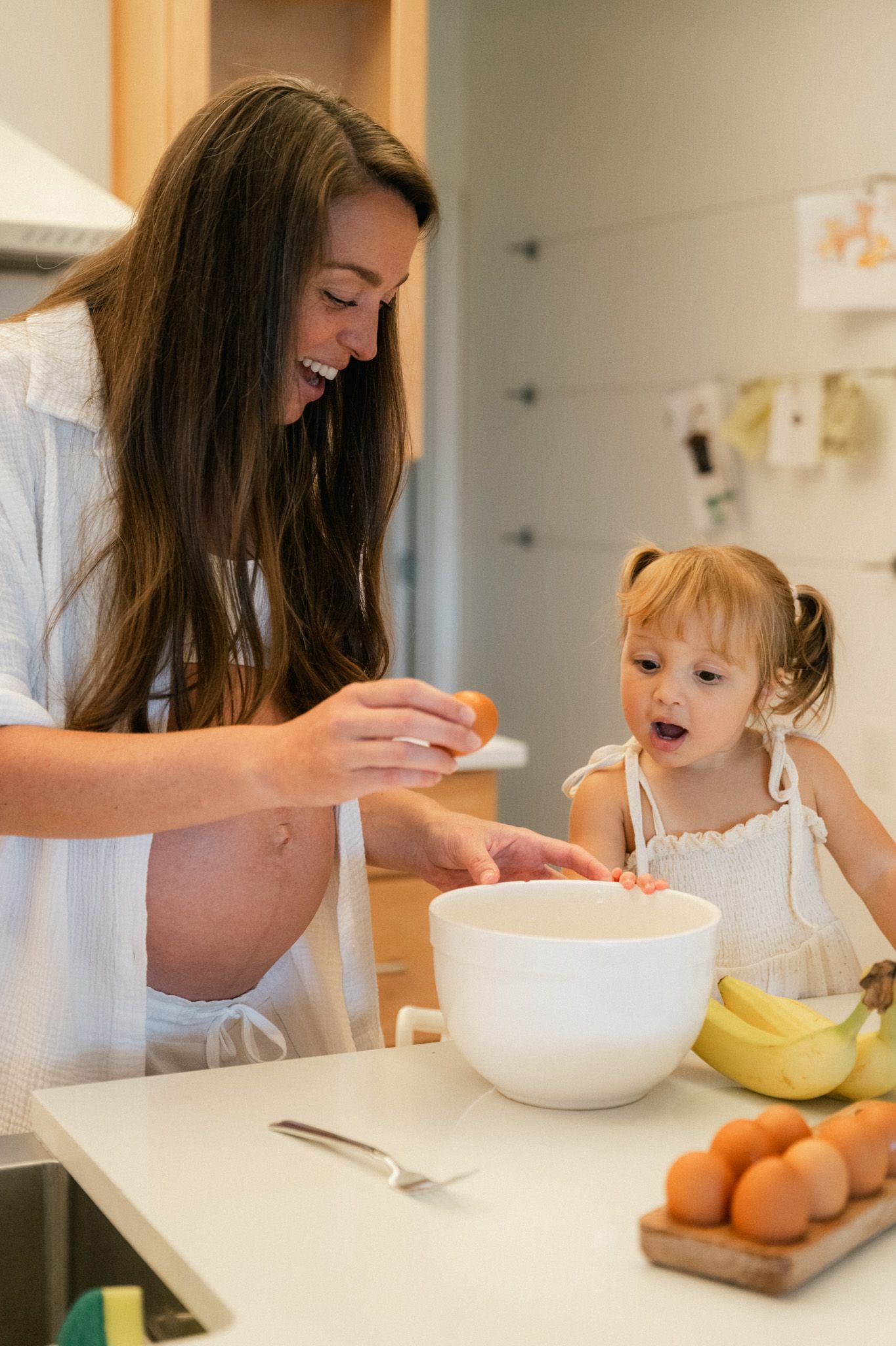 Pregnant woman and toddler girl cracking eggs into a bowl while baking together in a bright kitchen.