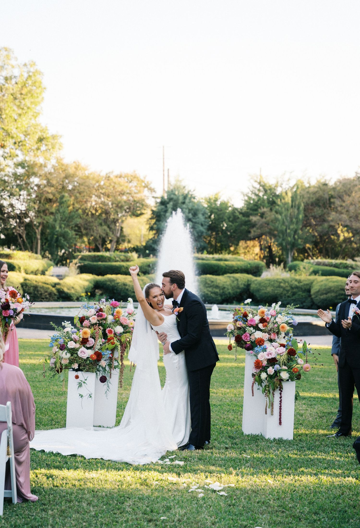 Newlyweds celebrate first kiss at outdoor wedding ceremony with colorful floral arrangements and fountain backdrop.