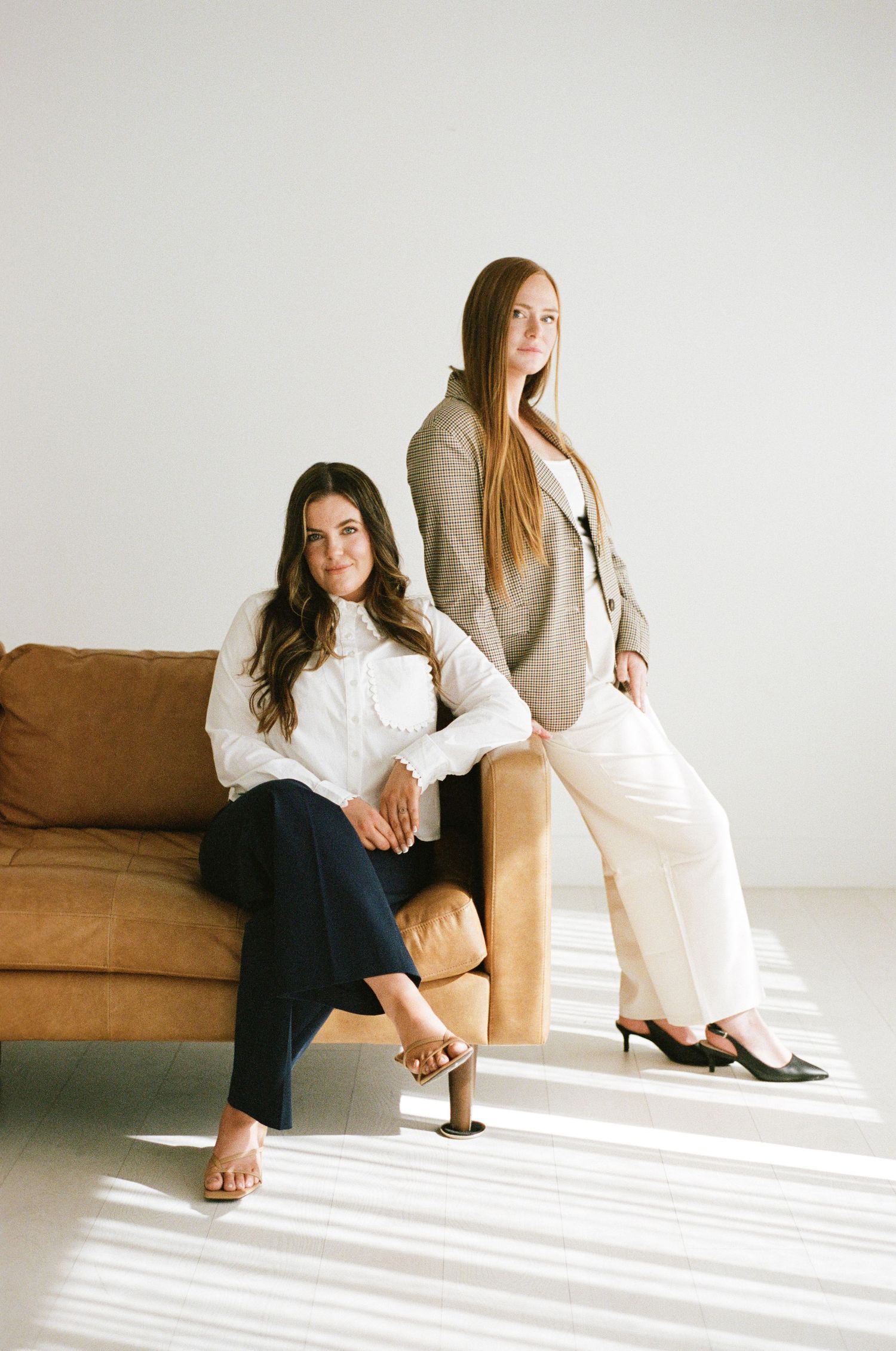 Two professional women posing by a tan leather sofa in a bright minimalist studio setting.