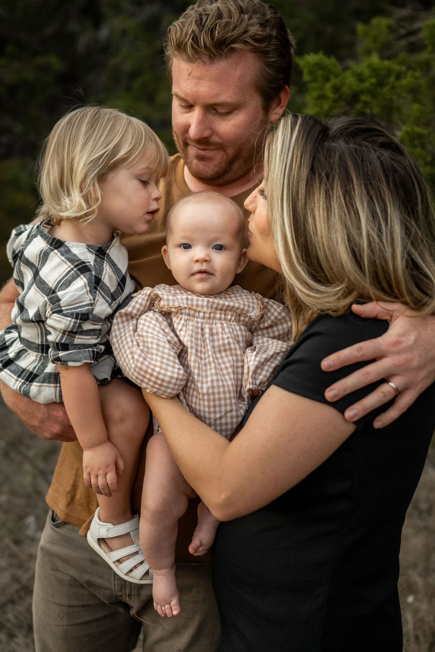 A young family with two small children share a tender moment outdoors, with parents holding their toddler and baby girl.