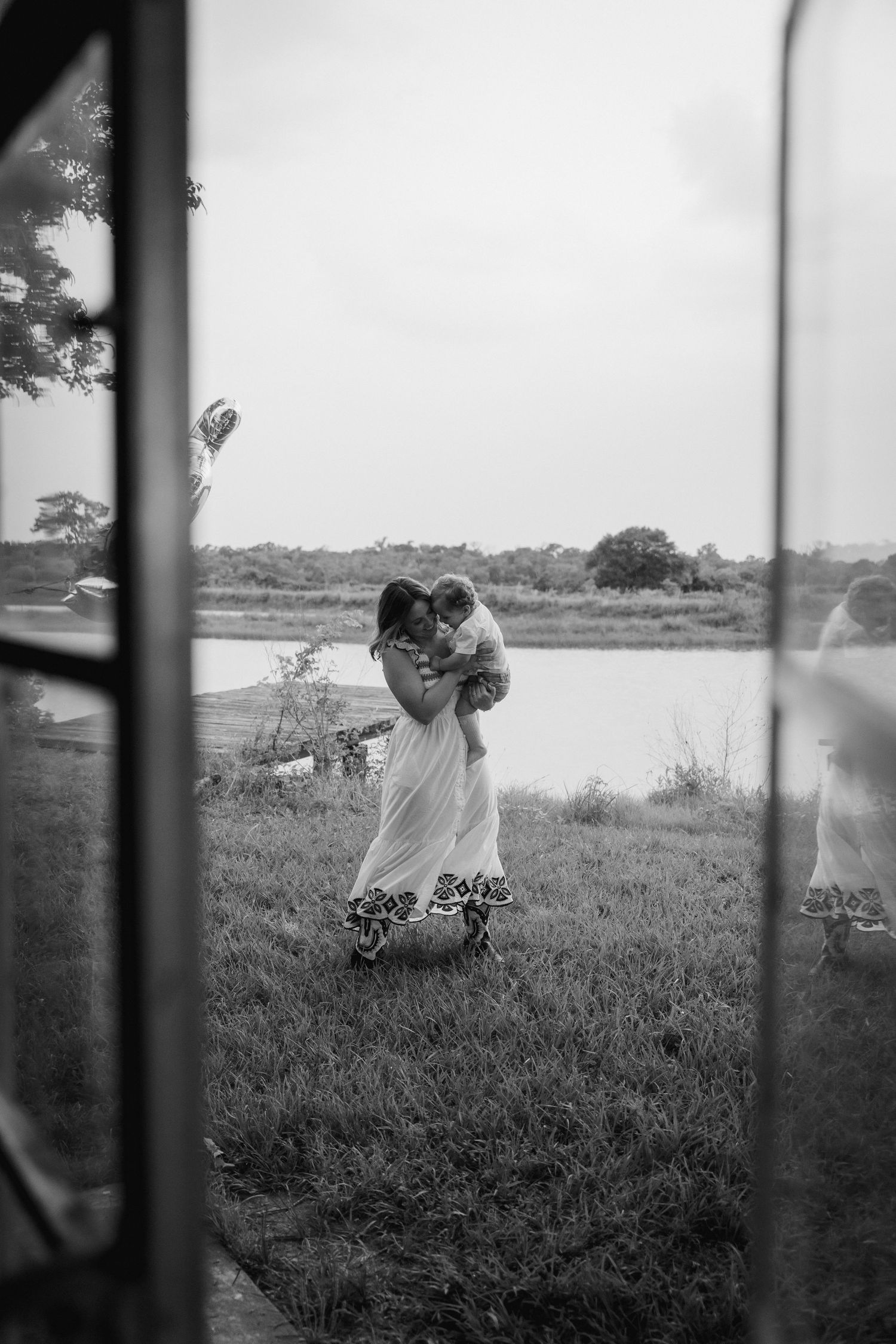 Black and white photo of a mother holding her child near a serene lake, viewed through a window frame.