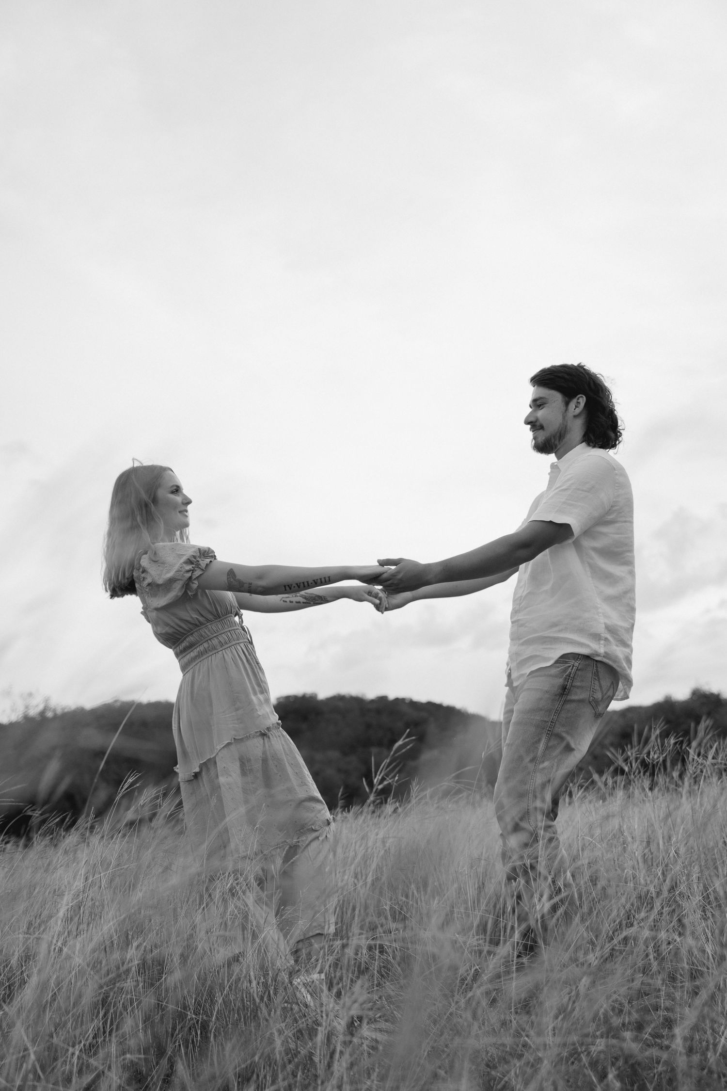 Black and white photo of a romantic couple holding hands in a grassy field, smiling at each other.