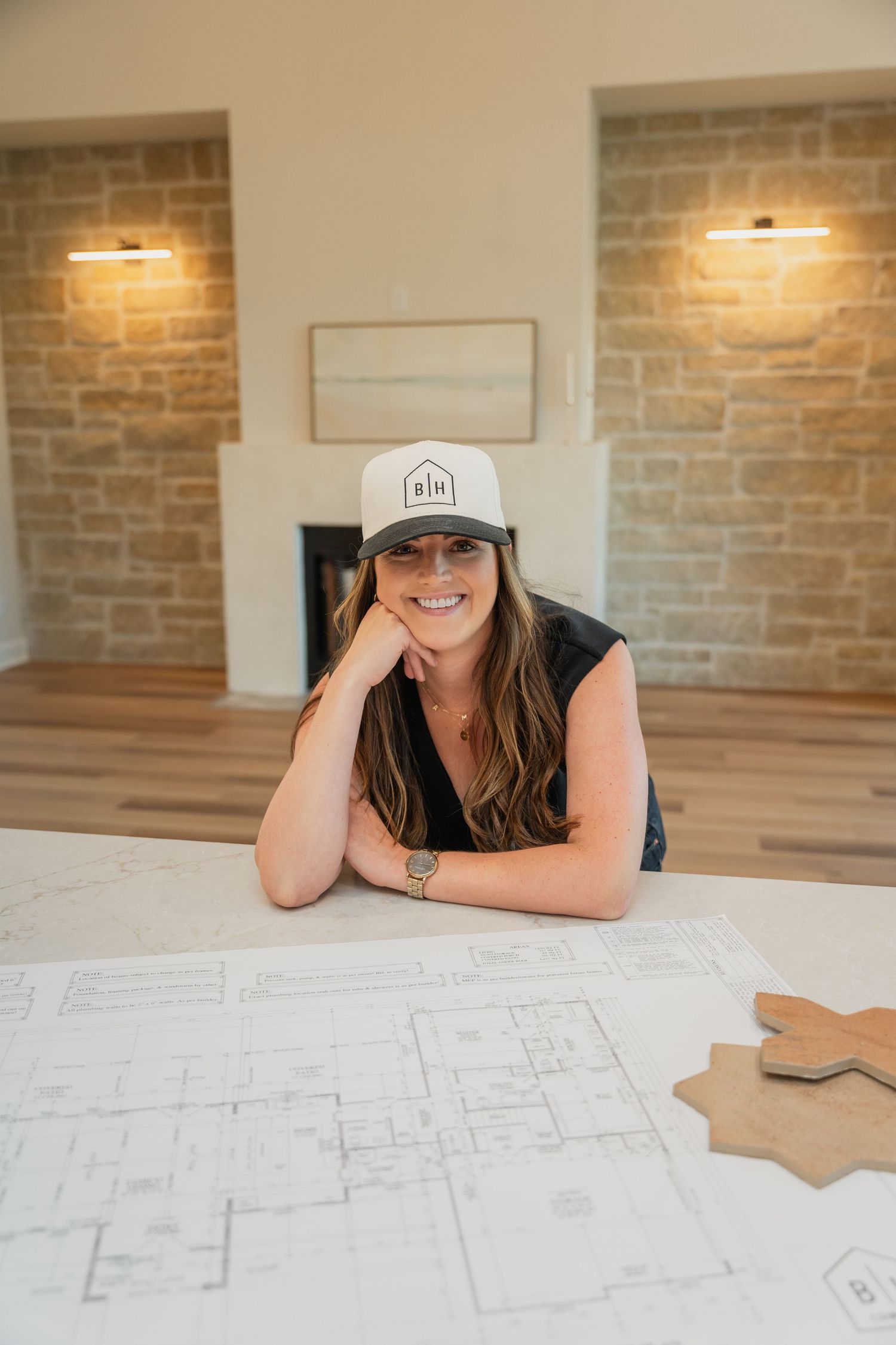 Woman in a cap smiles over architectural blueprints in a modern stone-walled interior.