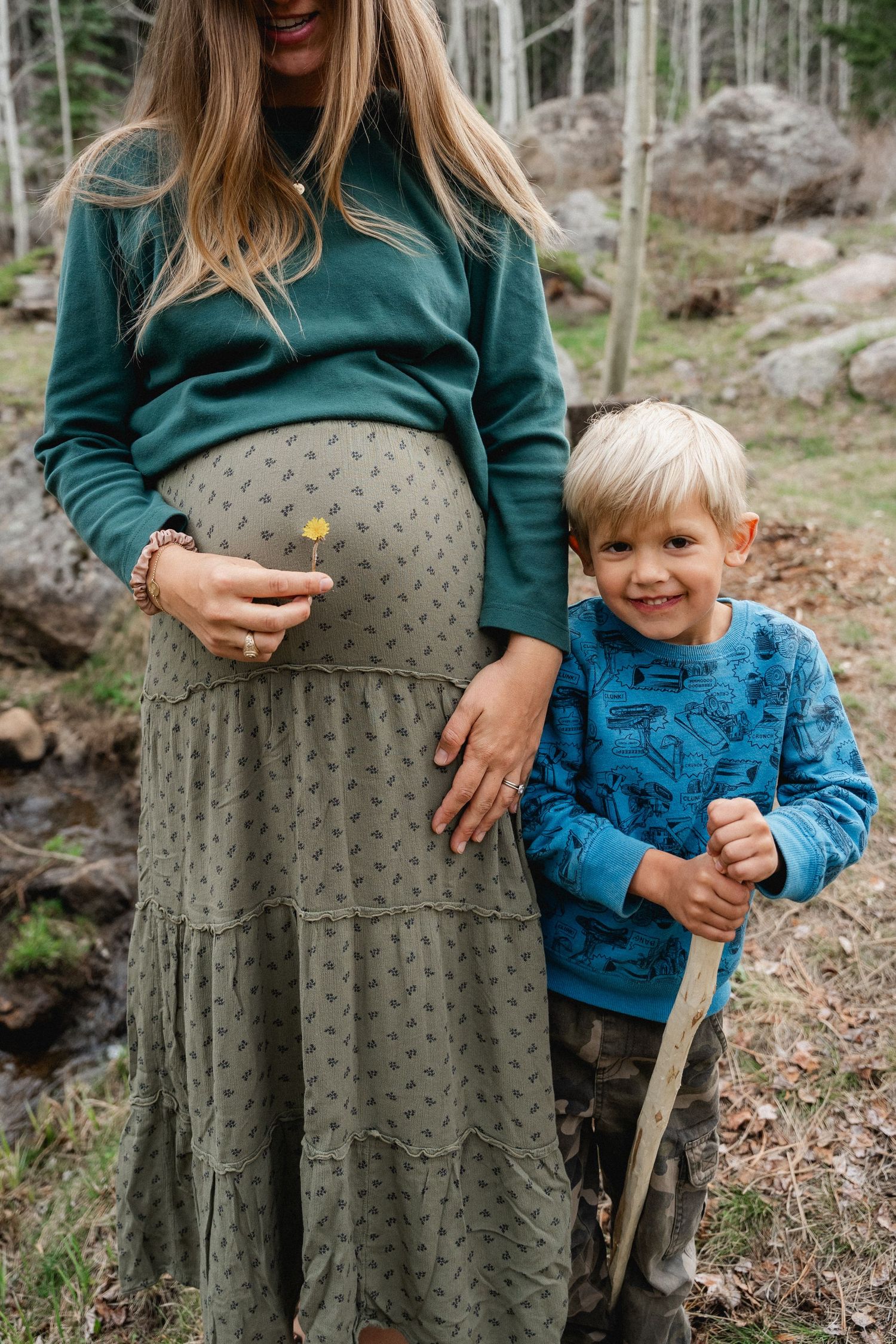 A pregnant woman in a green dress stands with a smiling blonde boy holding a stick in a forest.