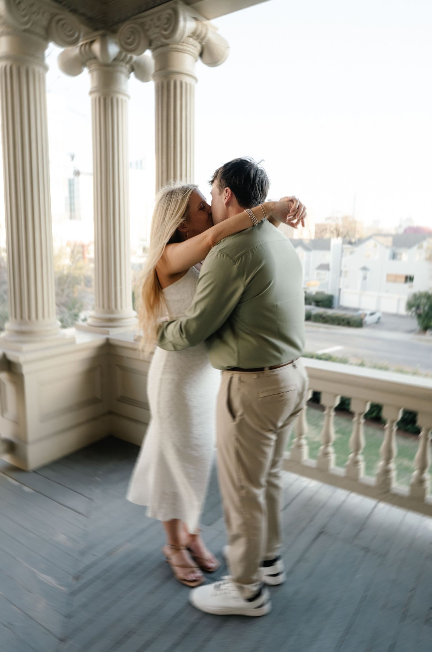 Couple sharing a romantic kiss on an elegant columned balcony with city views behind them.